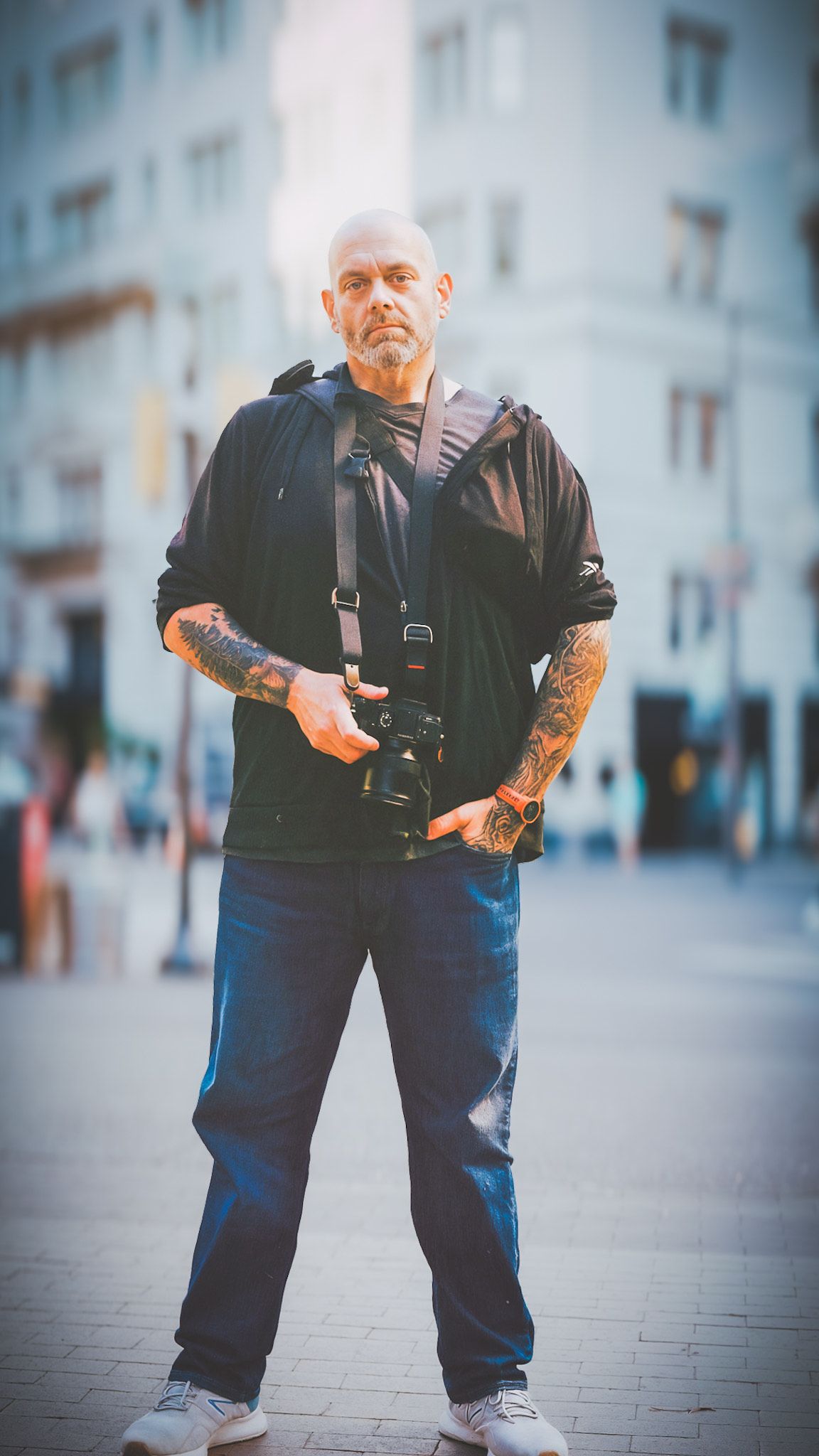 Person with tattoos wearing black jacket and jeans poses in urban setting with blurred background.