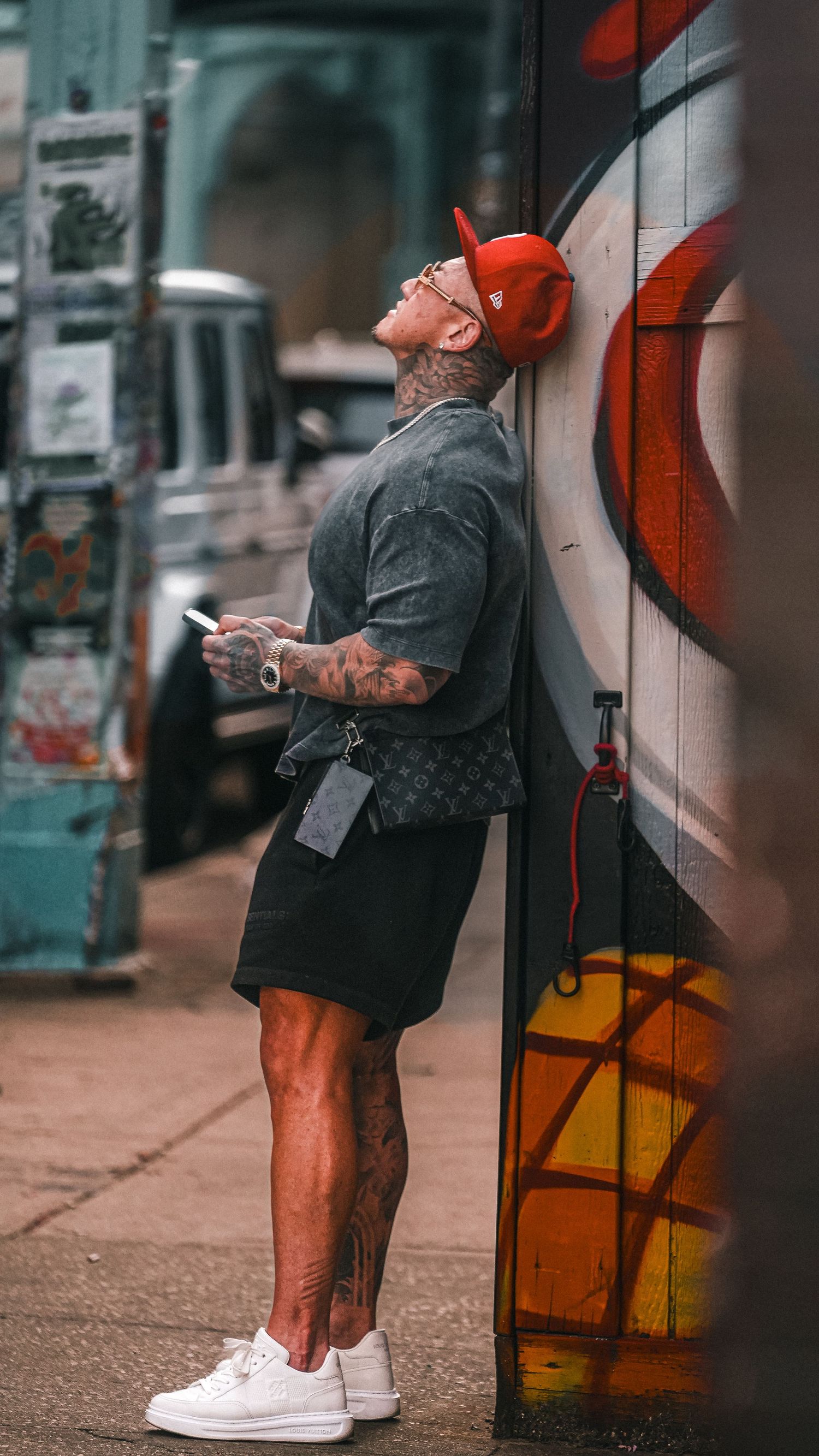 Person in red cap and shorts leans against graffiti wall while checking phone on urban street.