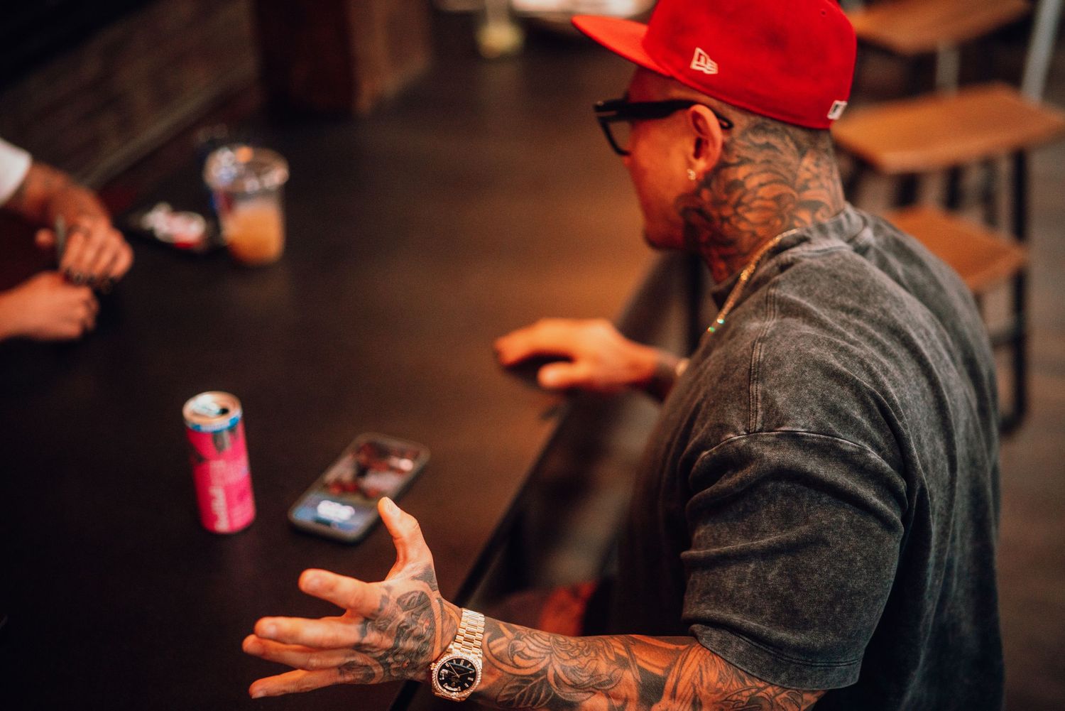 Person in red cap with tattoos sits at dark bar counter holding beverage while engaged in conversation.