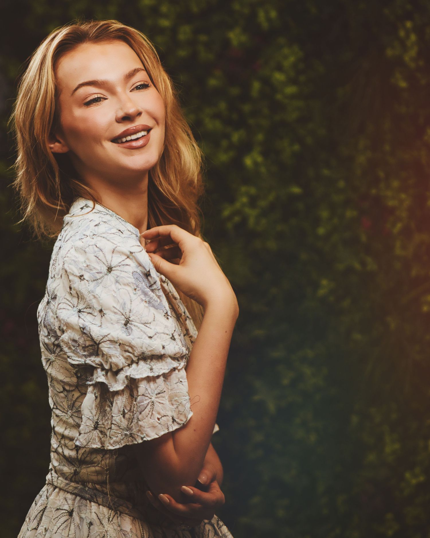Blonde woman in vintage-style lace dress poses joyfully against blurred natural background.