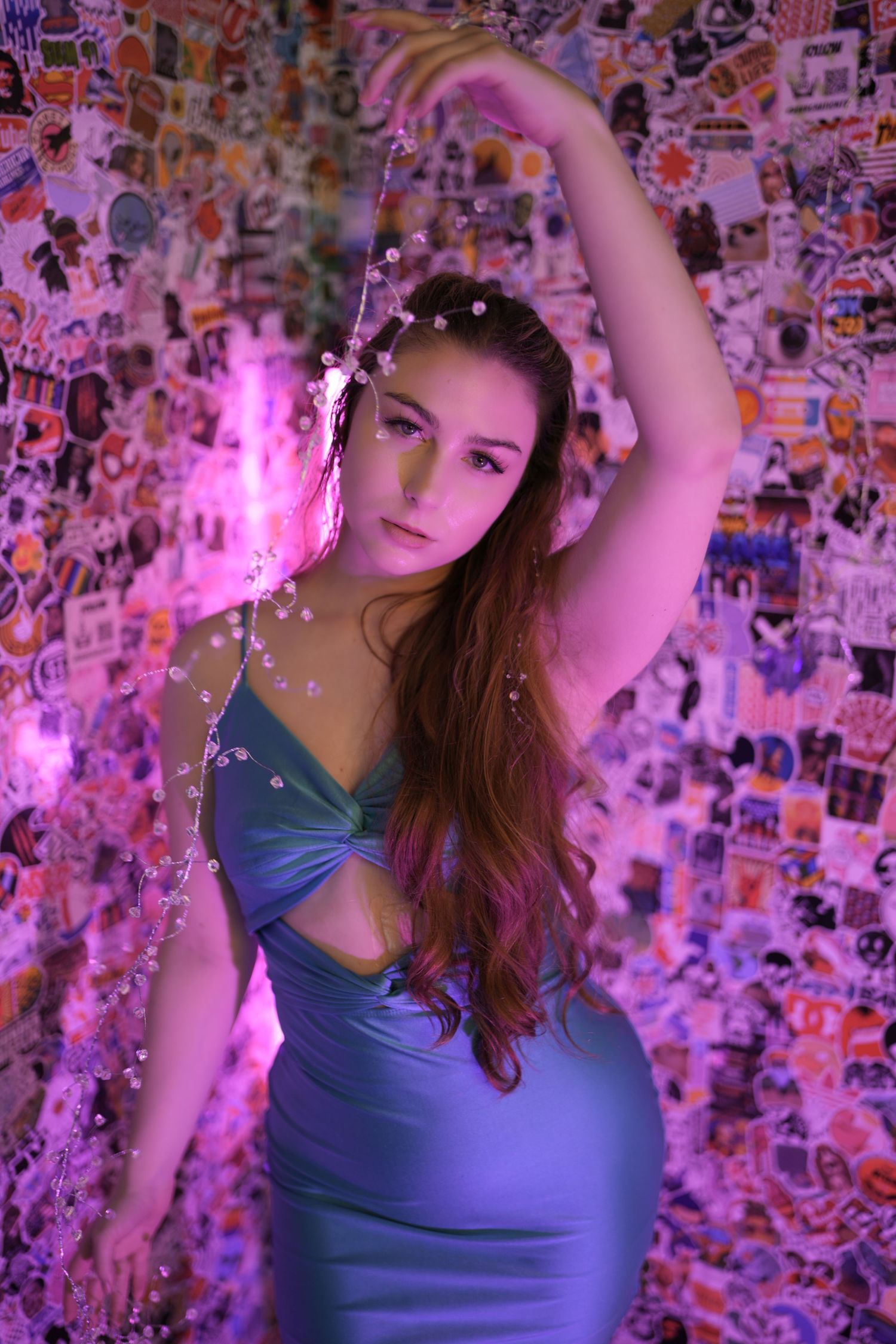 A figure in a blue tank top poses against a wall covered in colorful photos with pink lighting effects.