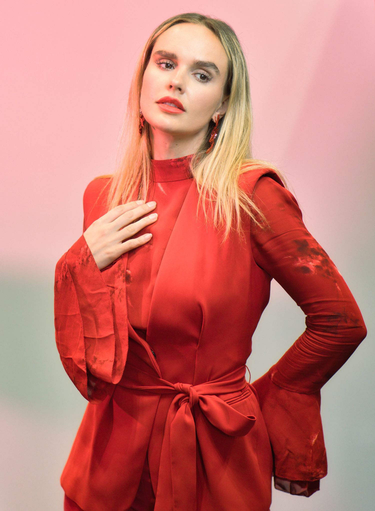 A high-fashion portrait in a vibrant red turtleneck dress with bell sleeves against a pink background.