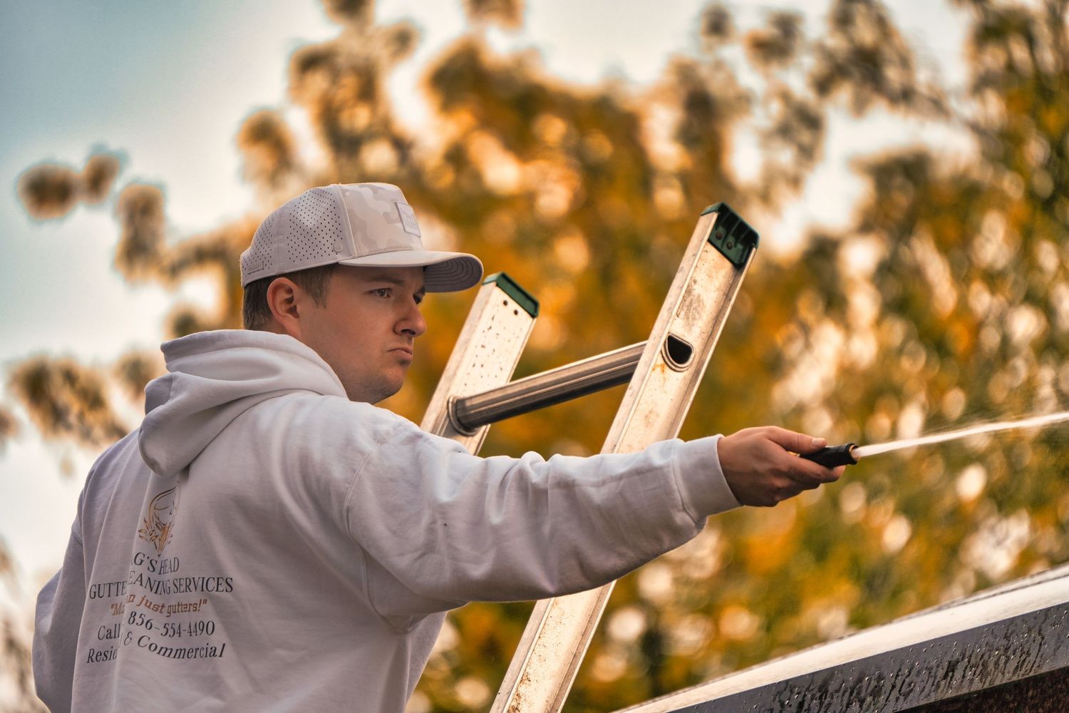 A person in a white hoodie and cap working on a ladder against an autumn background at sunset.
