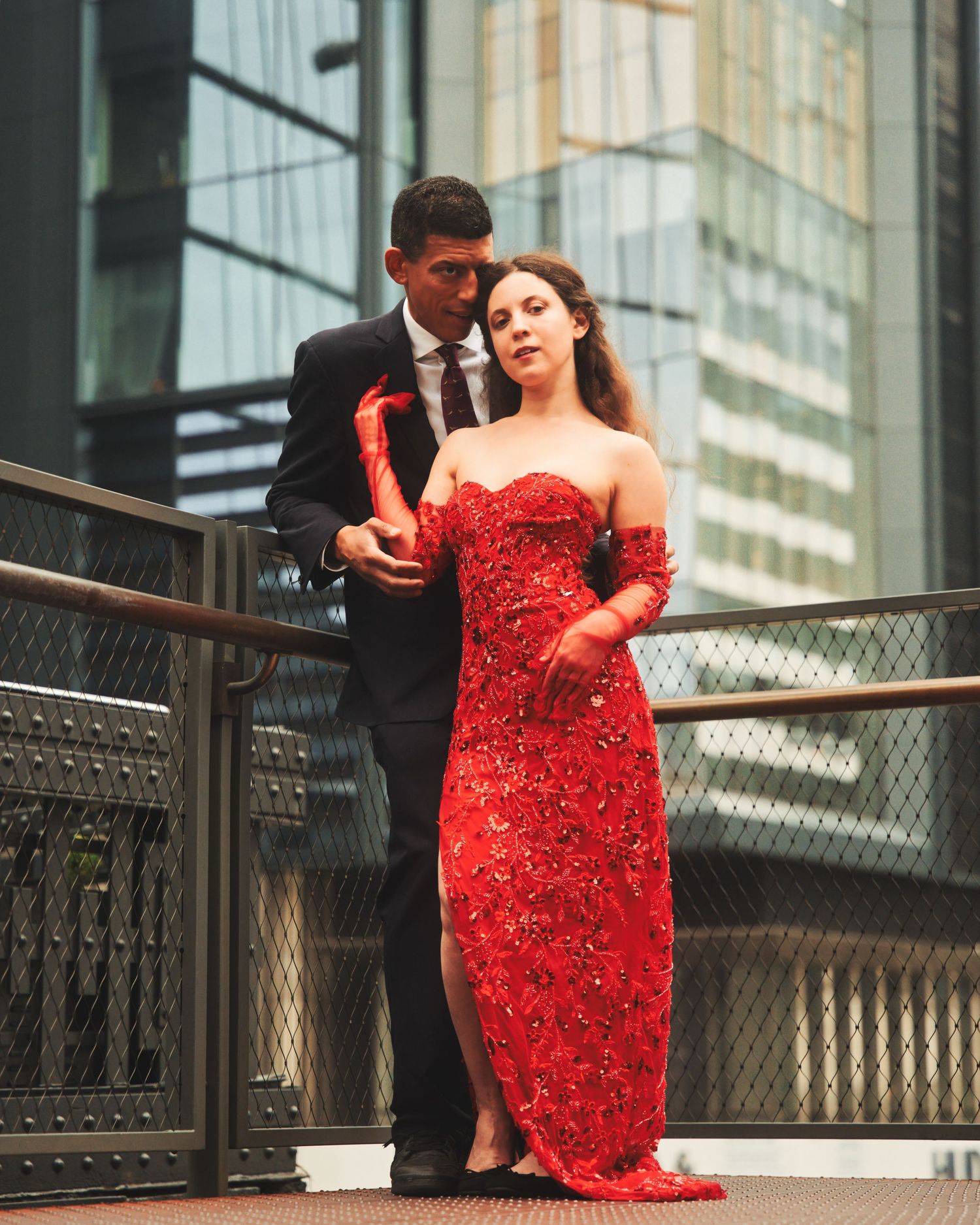A couple in formal attire pose on an urban walkway, with her wearing a stunning red sequined gown.