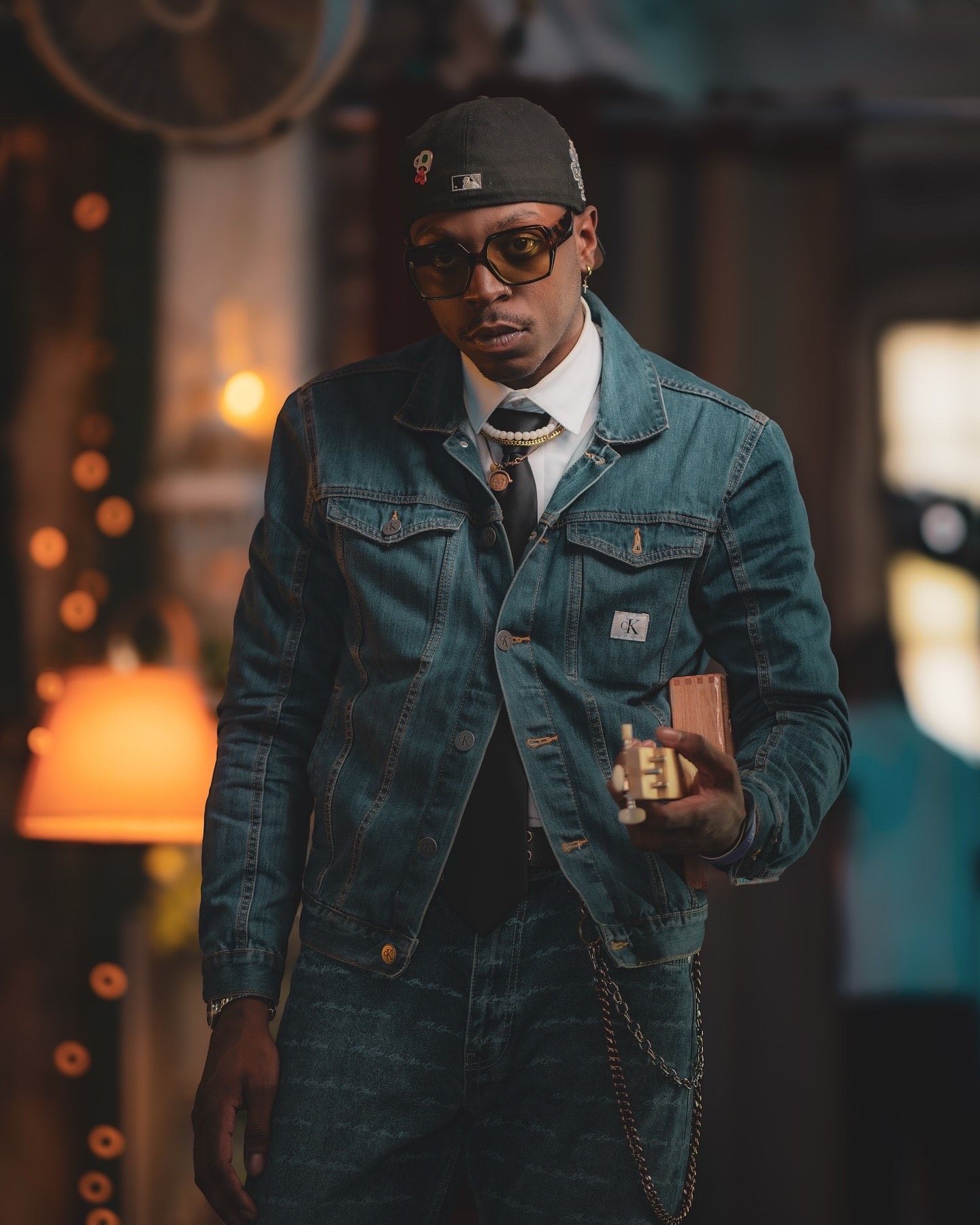 A well-dressed person in sunglasses and denim jacket walks through a dimly lit city setting with warm glowing lights.