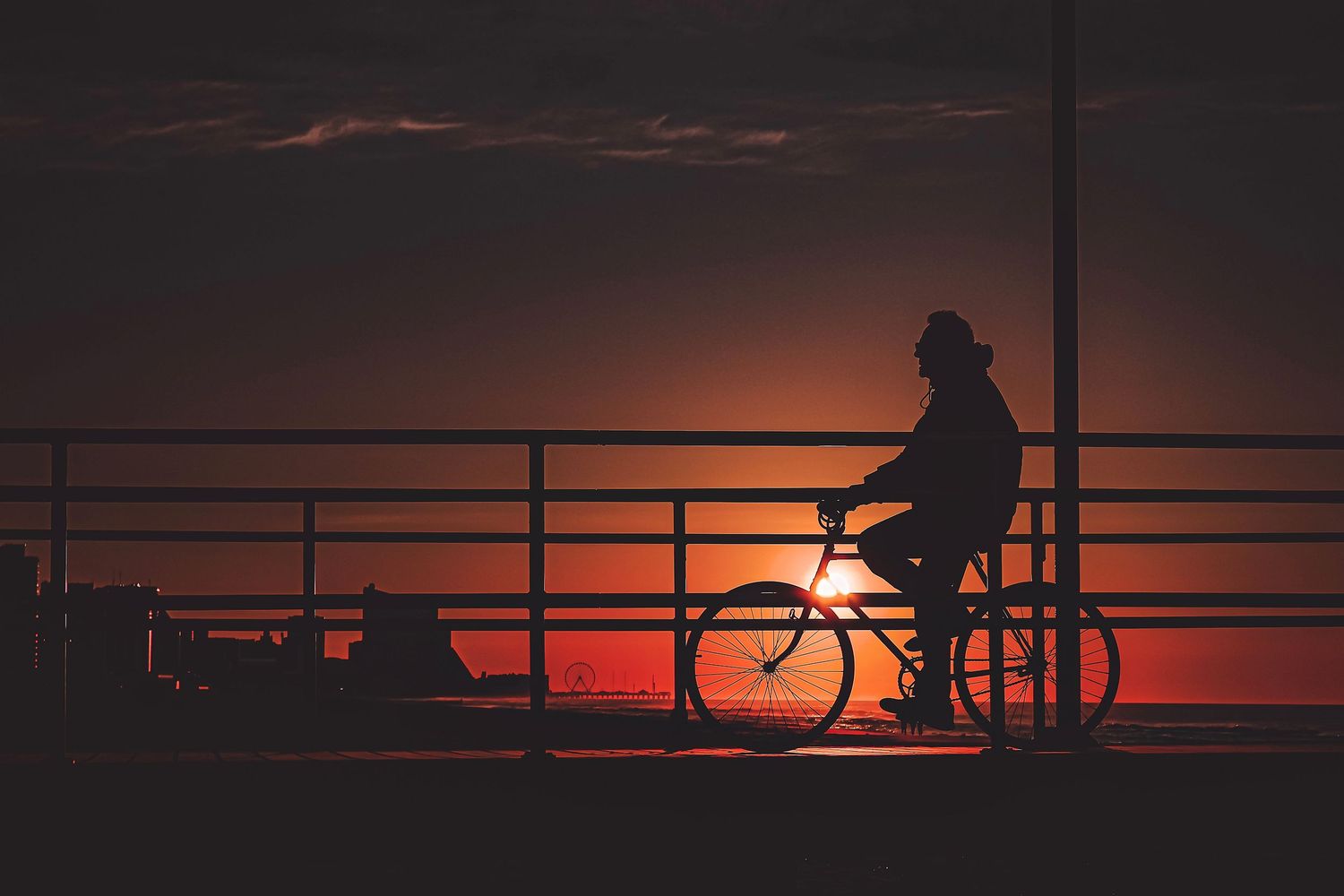 Silhouetted cyclist on a bridge railing against a vibrant orange sunset sky.