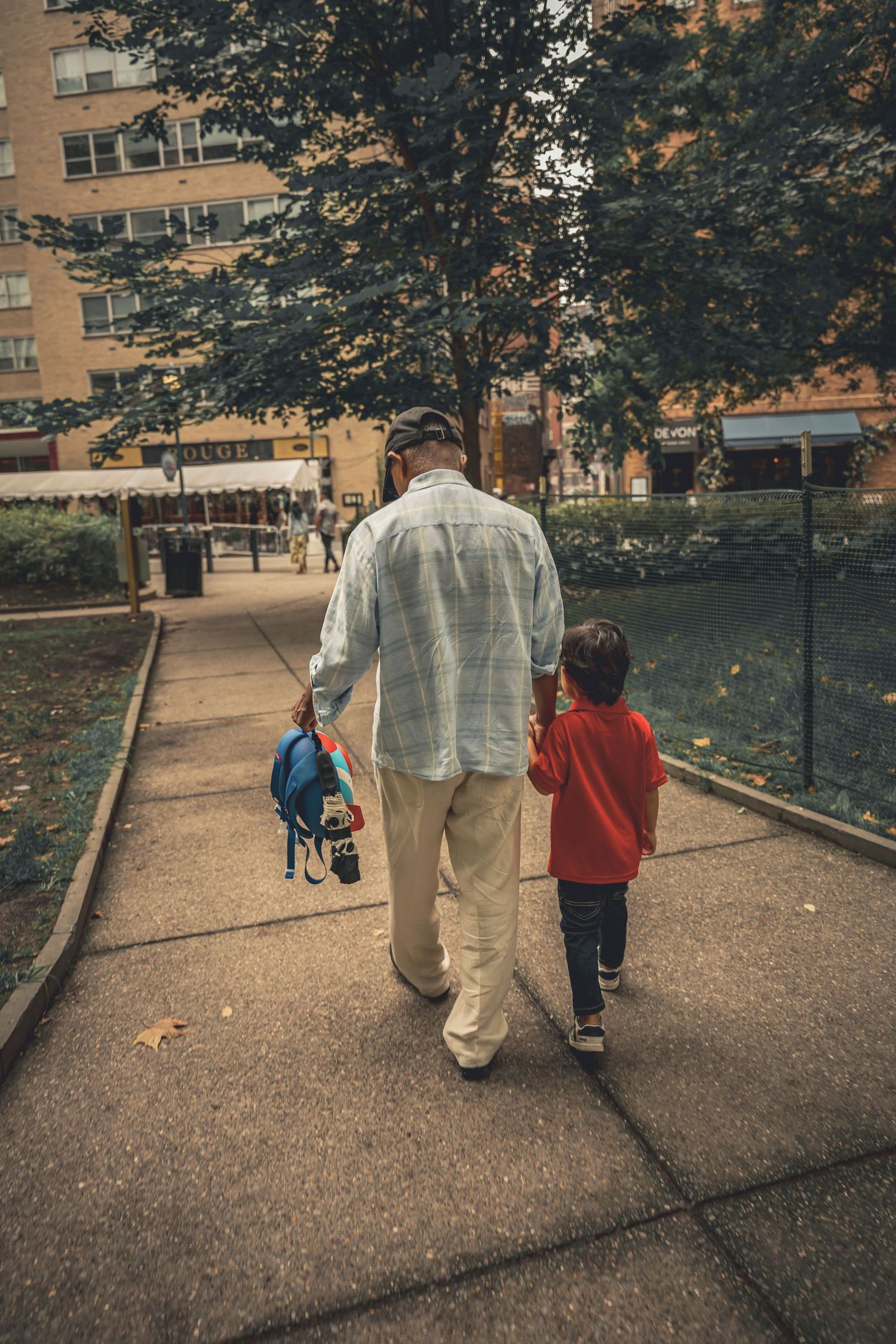 An adult and child walk together on a tree-lined path near apartment buildings on an autumn afternoon.
