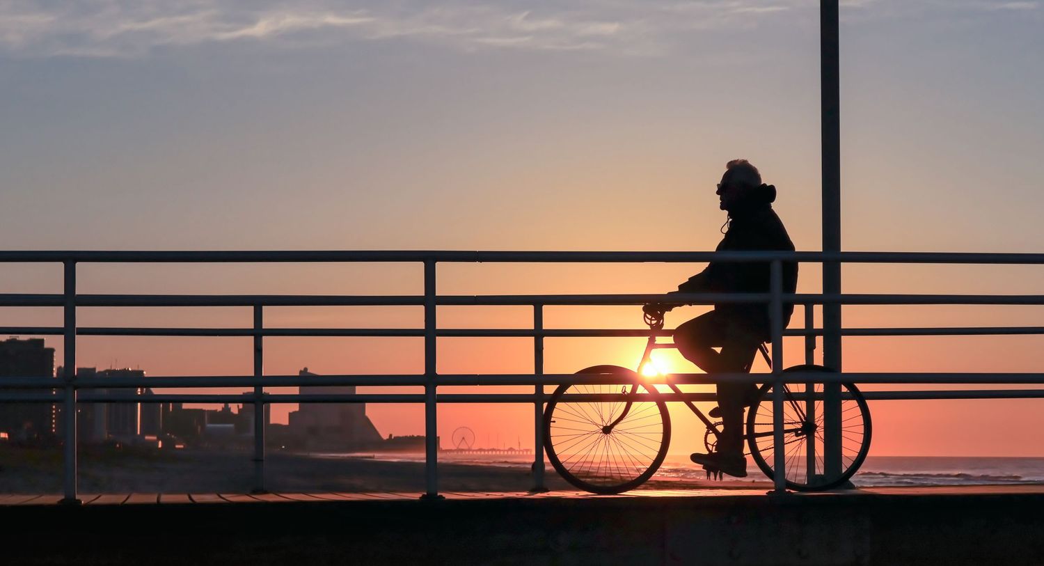 Silhouetted cyclists ride along a railing against a vibrant sunset sky over the water.