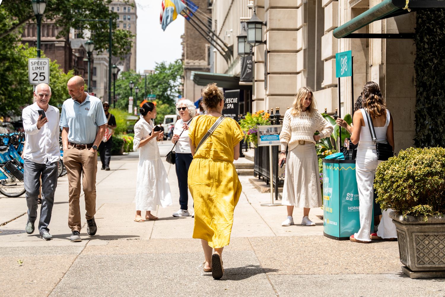 People walking down a busy urban sidewalk on a sunny day, with someone in a bright yellow dress standing out from the crowd.