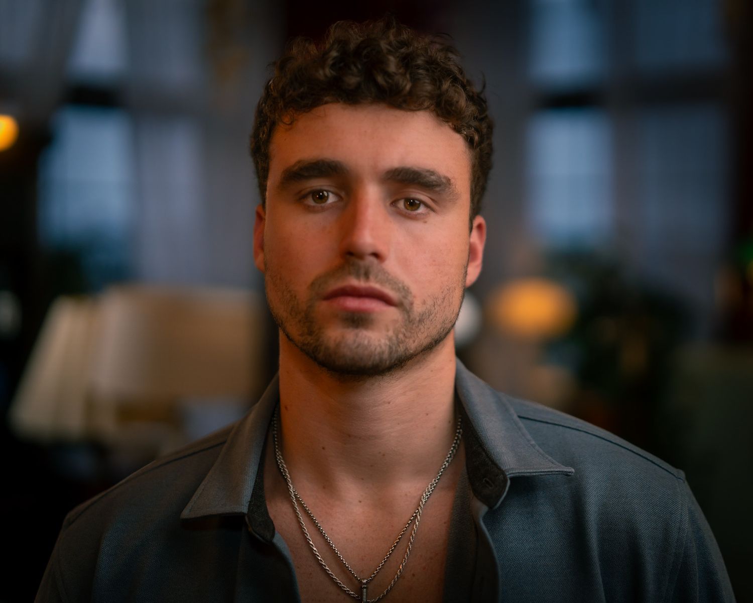 Portrait of a person with dark curly hair wearing a grey button-up shirt and necklace in moody indoor lighting.