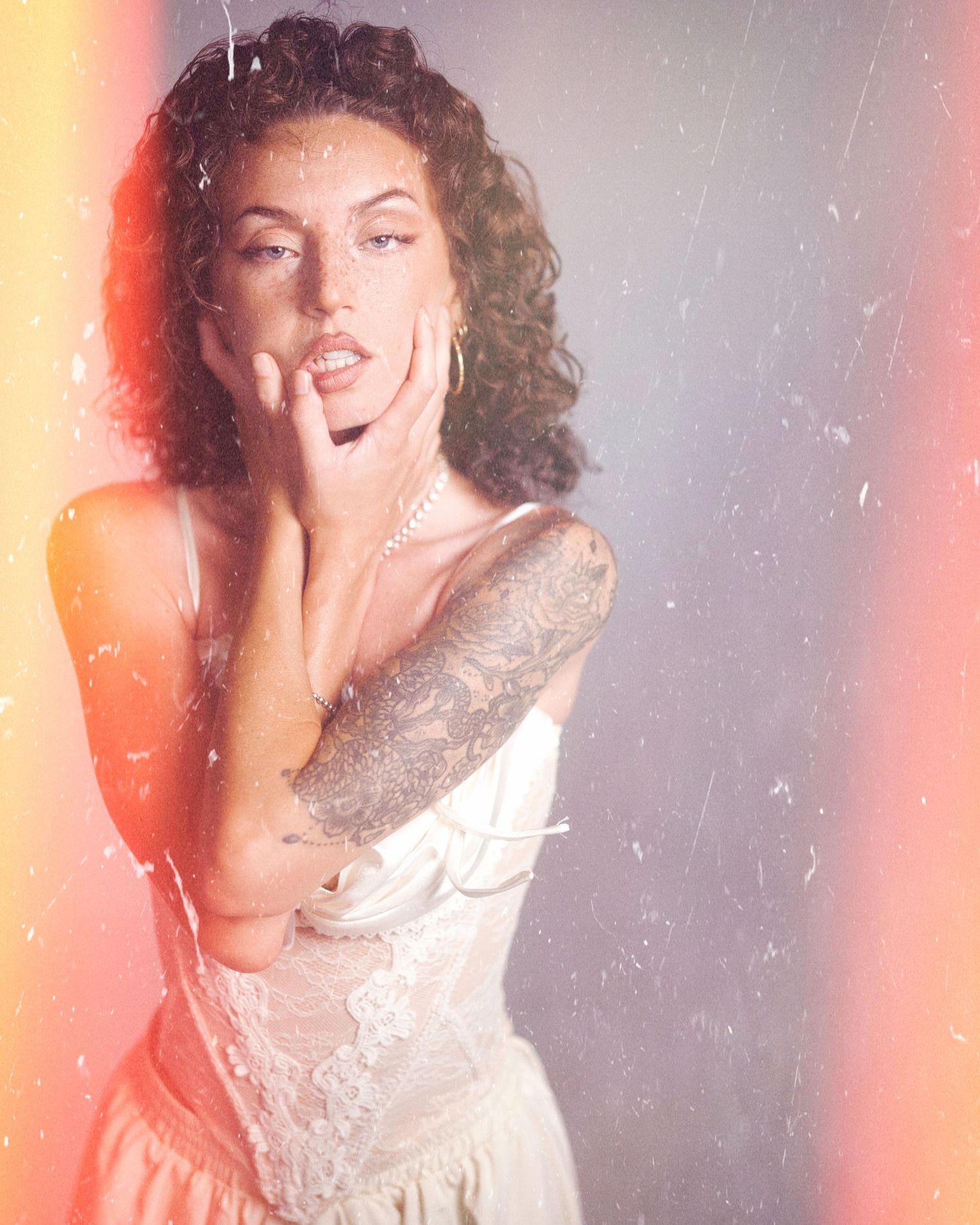 A person in a vintage-style white lace dress poses dramatically with hand to face against a light background with pink glow.