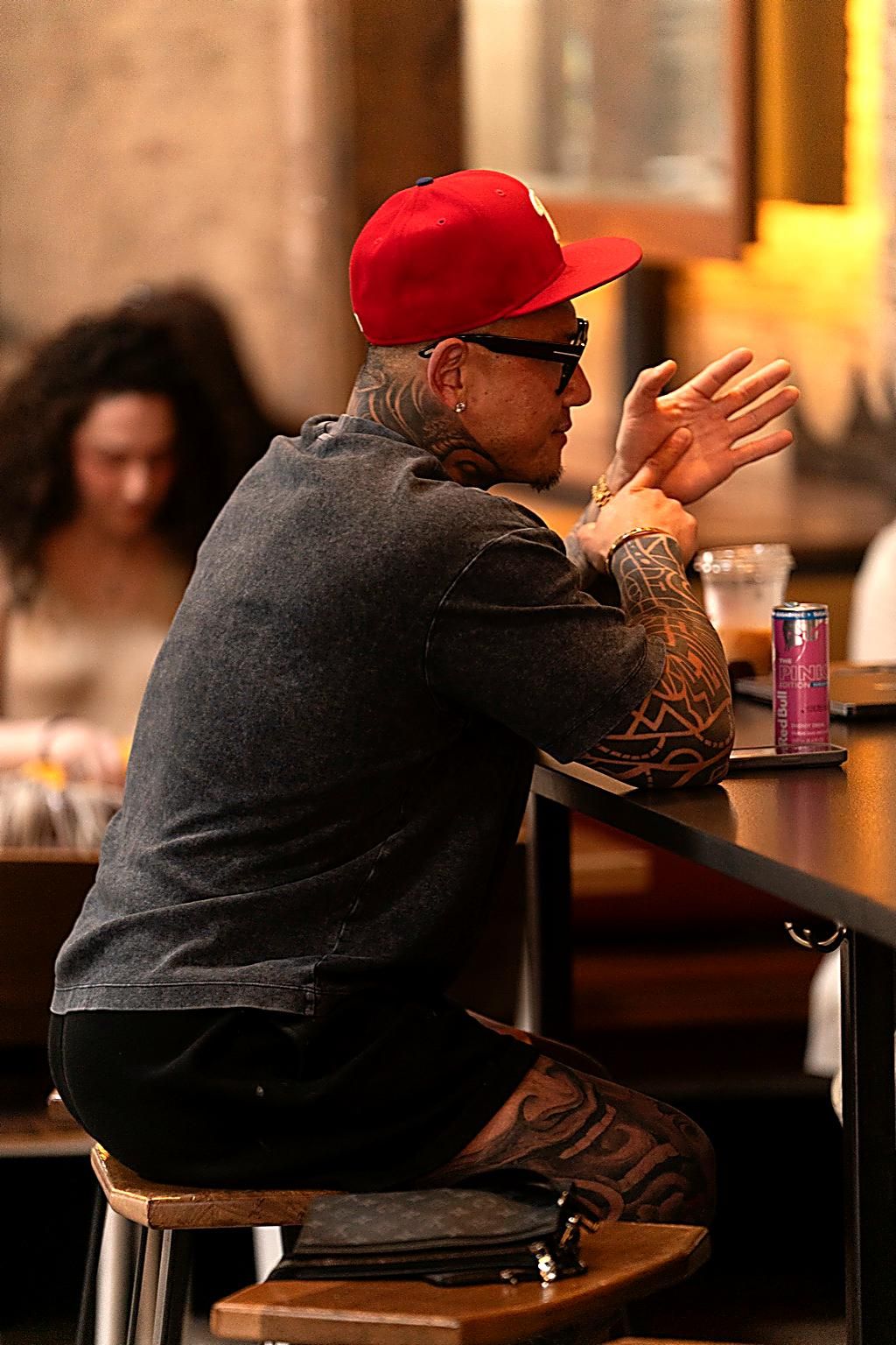 A person in a red cap and dark jacket with tattoos sits at a bar counter gesturing while talking.