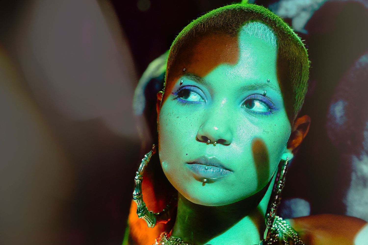 Figure illuminated by dramatic green lighting wears ornate earrings and an orange scarf against a dark background.