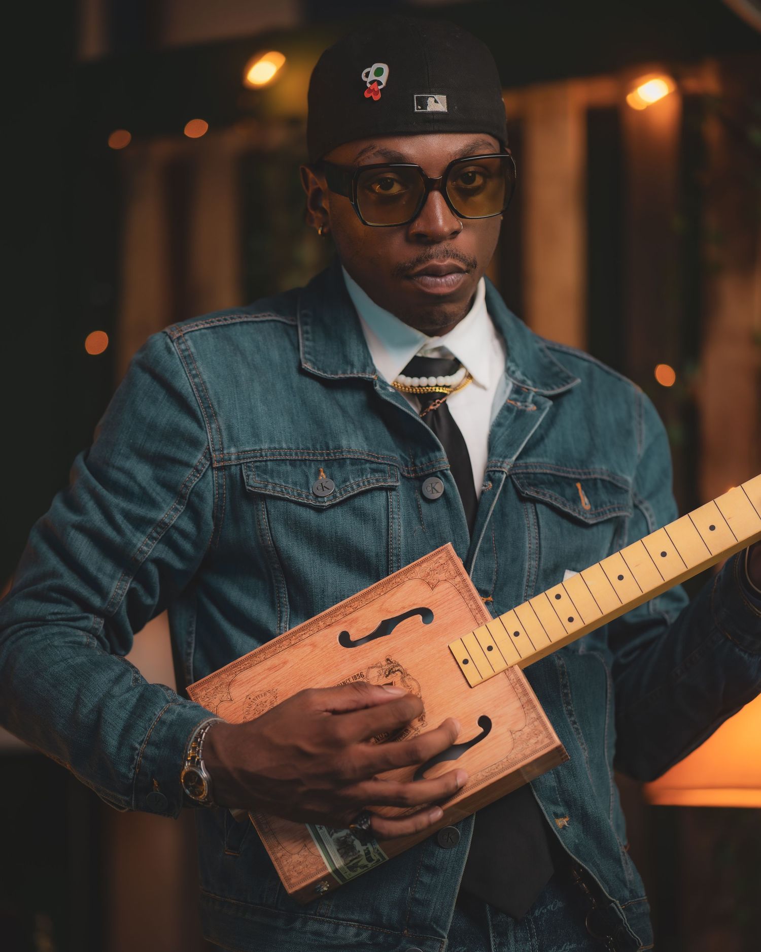 Musician in denim jacket and baseball cap holding a handcrafted wooden guitar in moody lighting.
