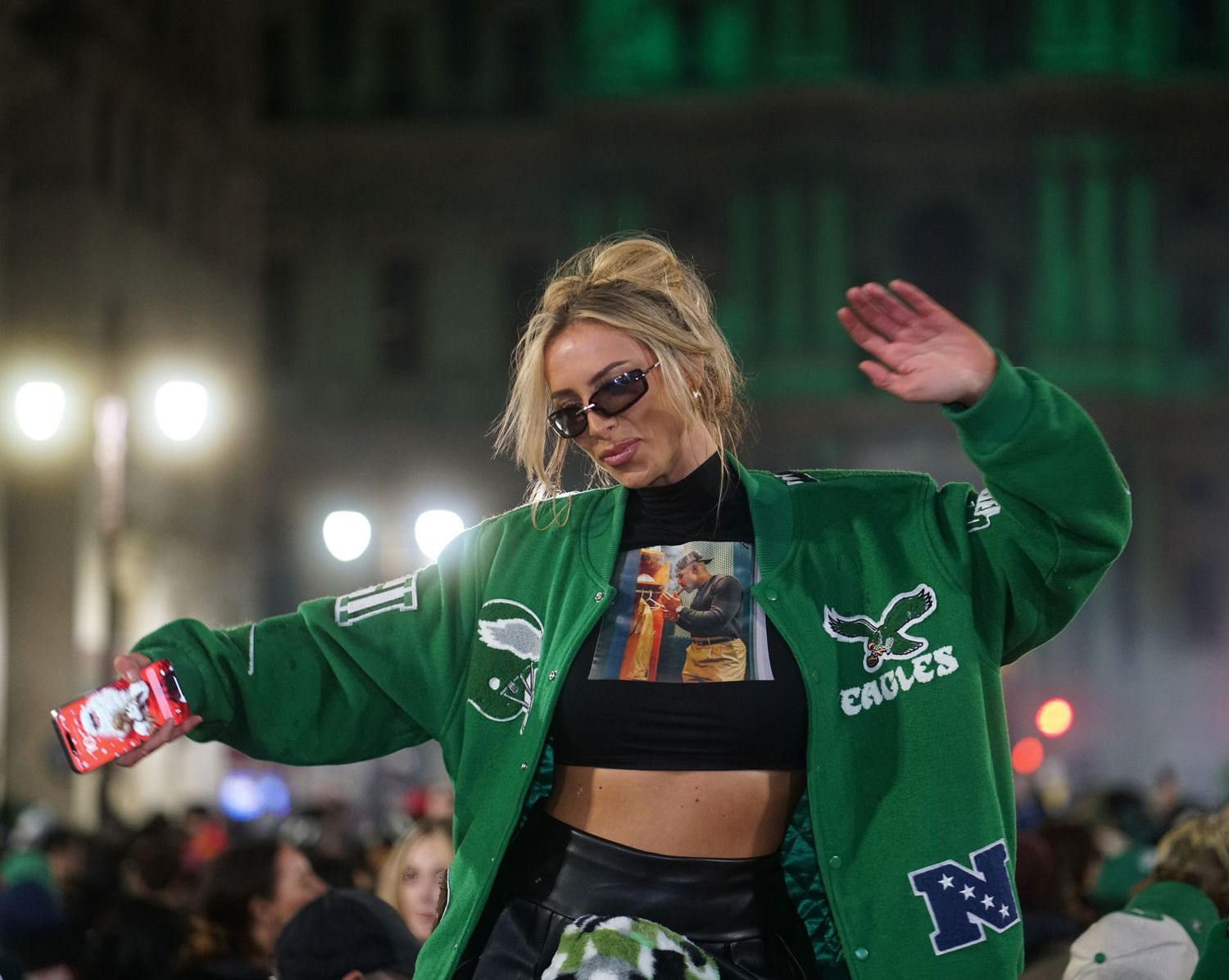 A person in a green varsity jacket and sunglasses waves while holding a can at a nighttime outdoor event.