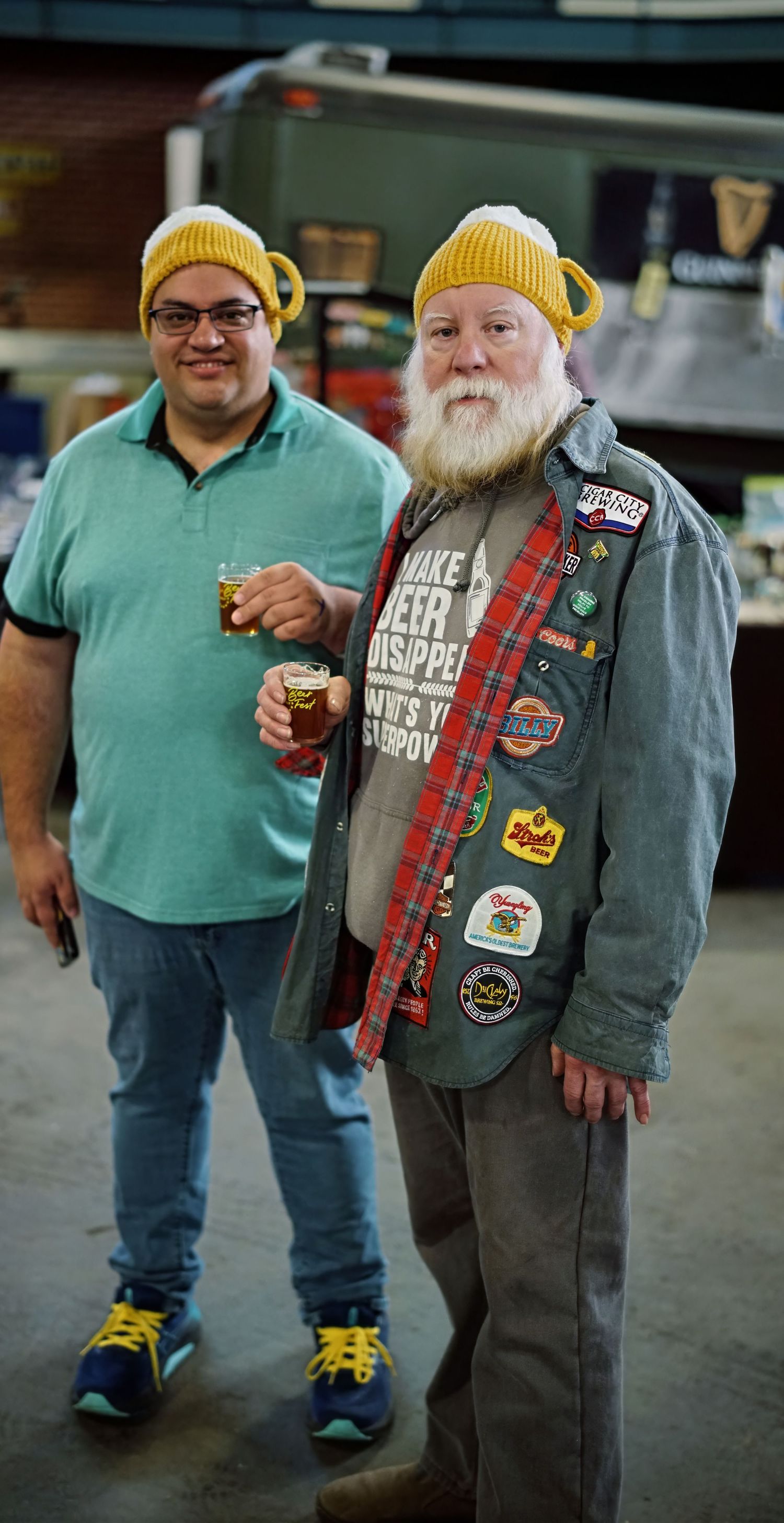 Two beer enthusiasts wearing yellow beanies stand together, with one sporting a denim jacket covered in brewery patches.
