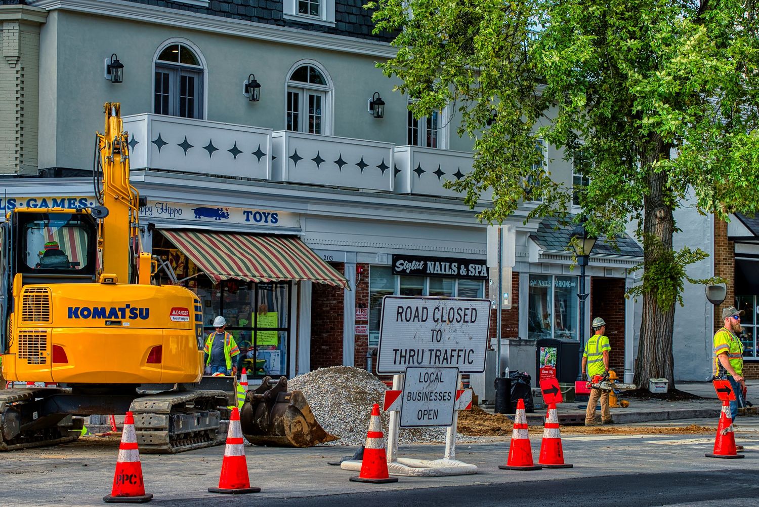 Construction equipment and safety cones block off a section of road in front of historic white buildings in a downtown area.