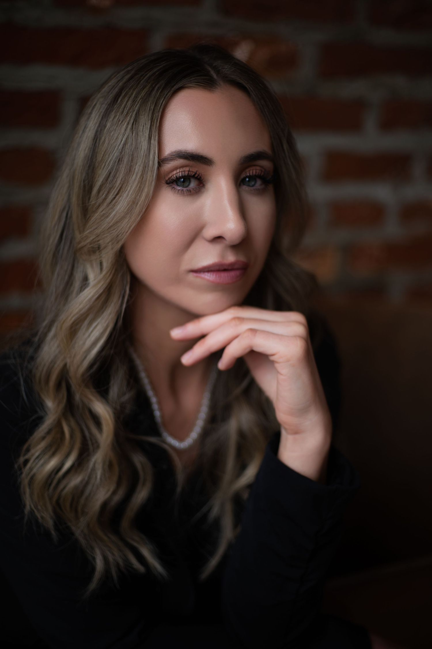 Portrait of someone with wavy blonde hair and a black top posing thoughtfully against a brick wall in moody lighting.