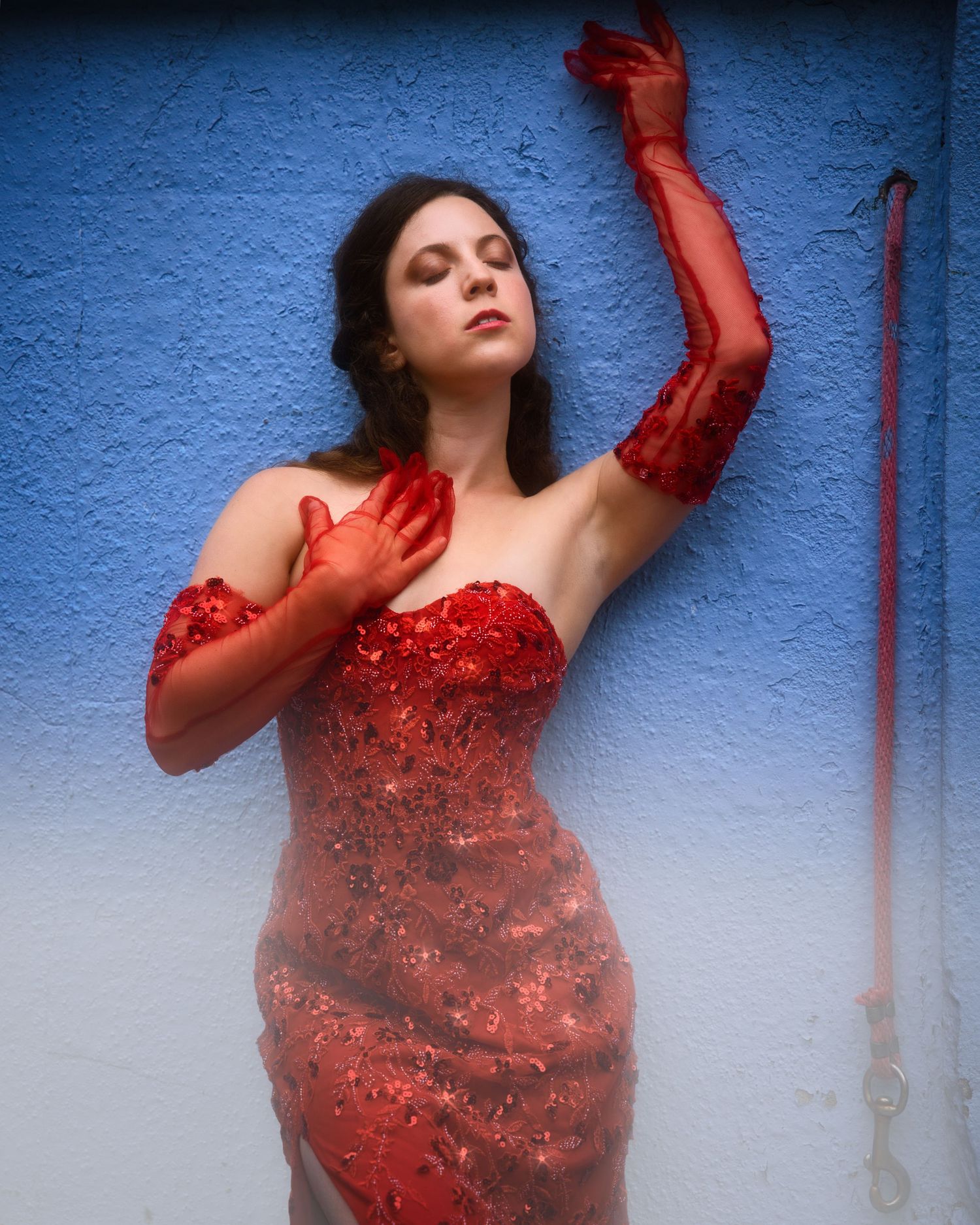 Model Katriella Dramatic fashion sequence of a figure in a sparkling red dress posing artistically against a textured blue wall.