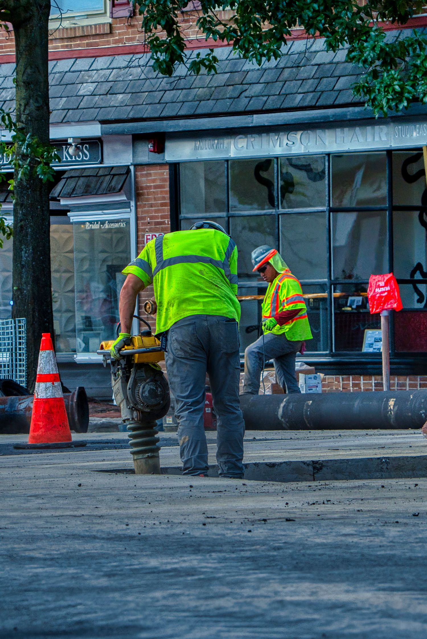 Construction workers in high-visibility gear operating equipment on a street with orange safety cones near a storefront.