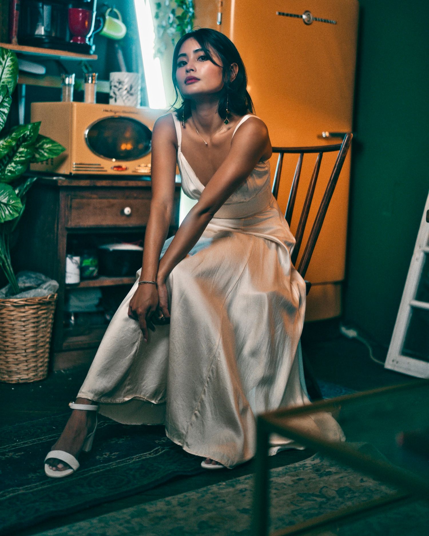 A dreamy indoor portrait shows someone in an elegant satin dress sitting on a wooden chair in a vintage-styled room.