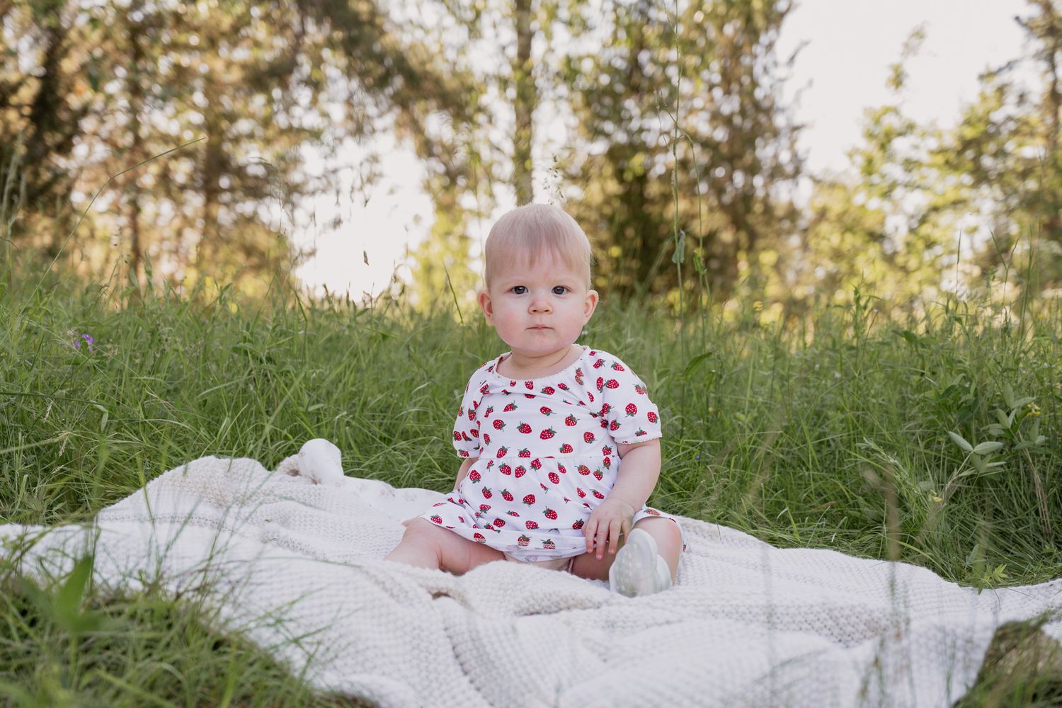 Baby in cherry-print onesie sitting on white blanket in green grassy field with trees in background.