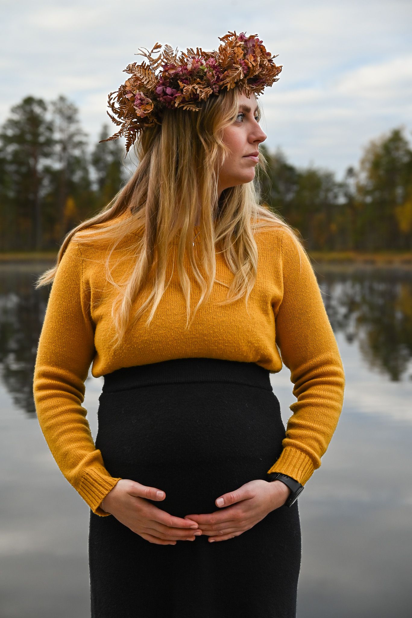 Gravid kvinna i gul stickad tröja med höstig blomkrans i håret framför tjärn en tidig höstmorgon.