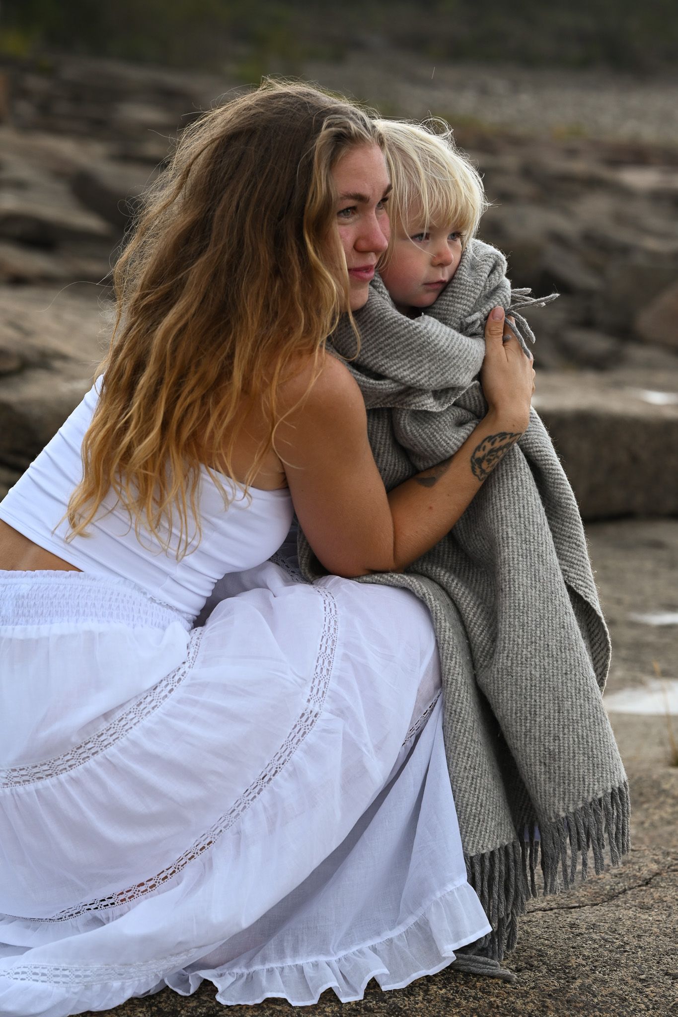 Kärleksfullt ögonblick på stranden intill havet med barn insvept i mysig filt och mamma i boho-klänning.