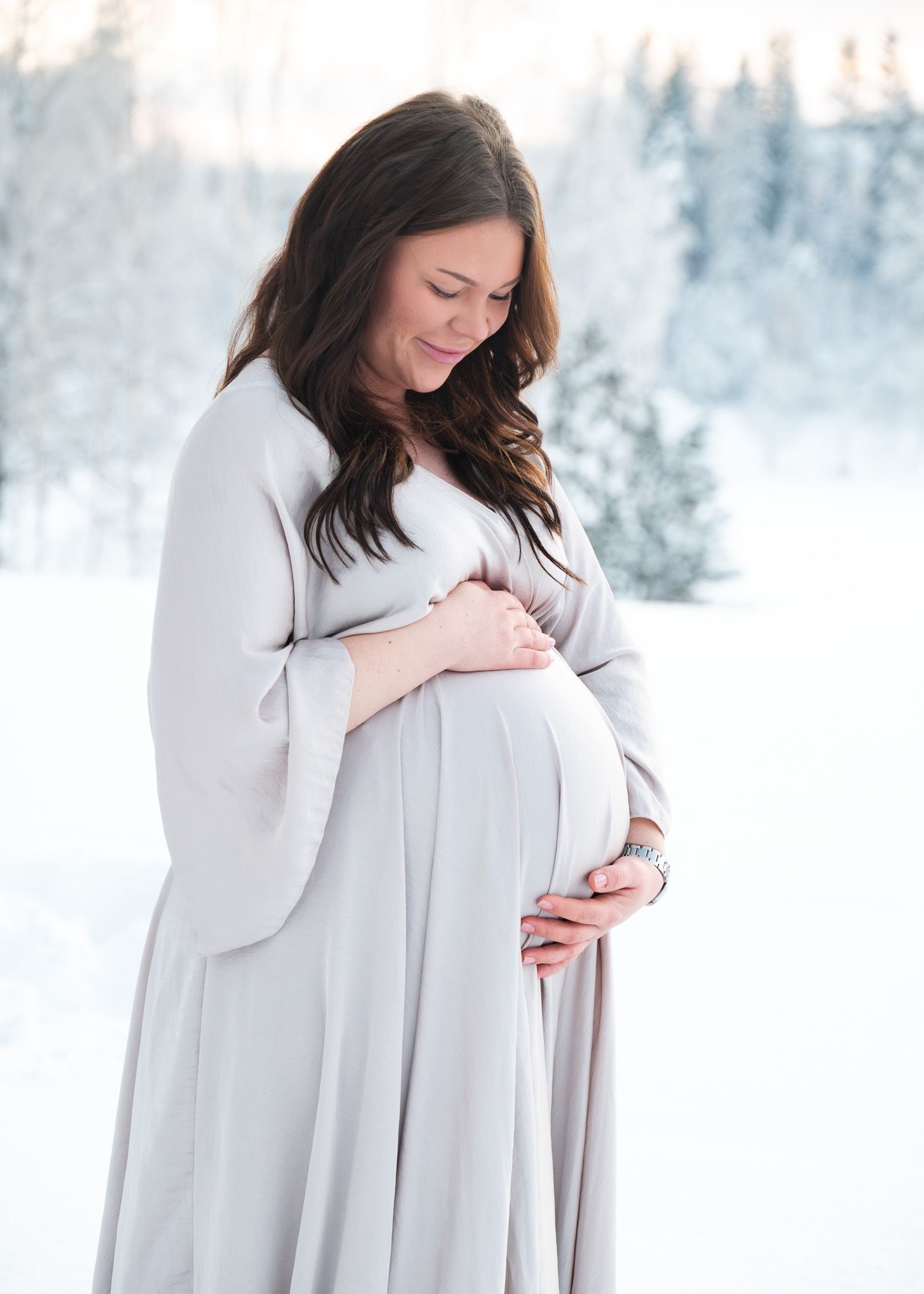 Gravid kvinna i beige klänning under gravidfotografering i snön och golden hour.