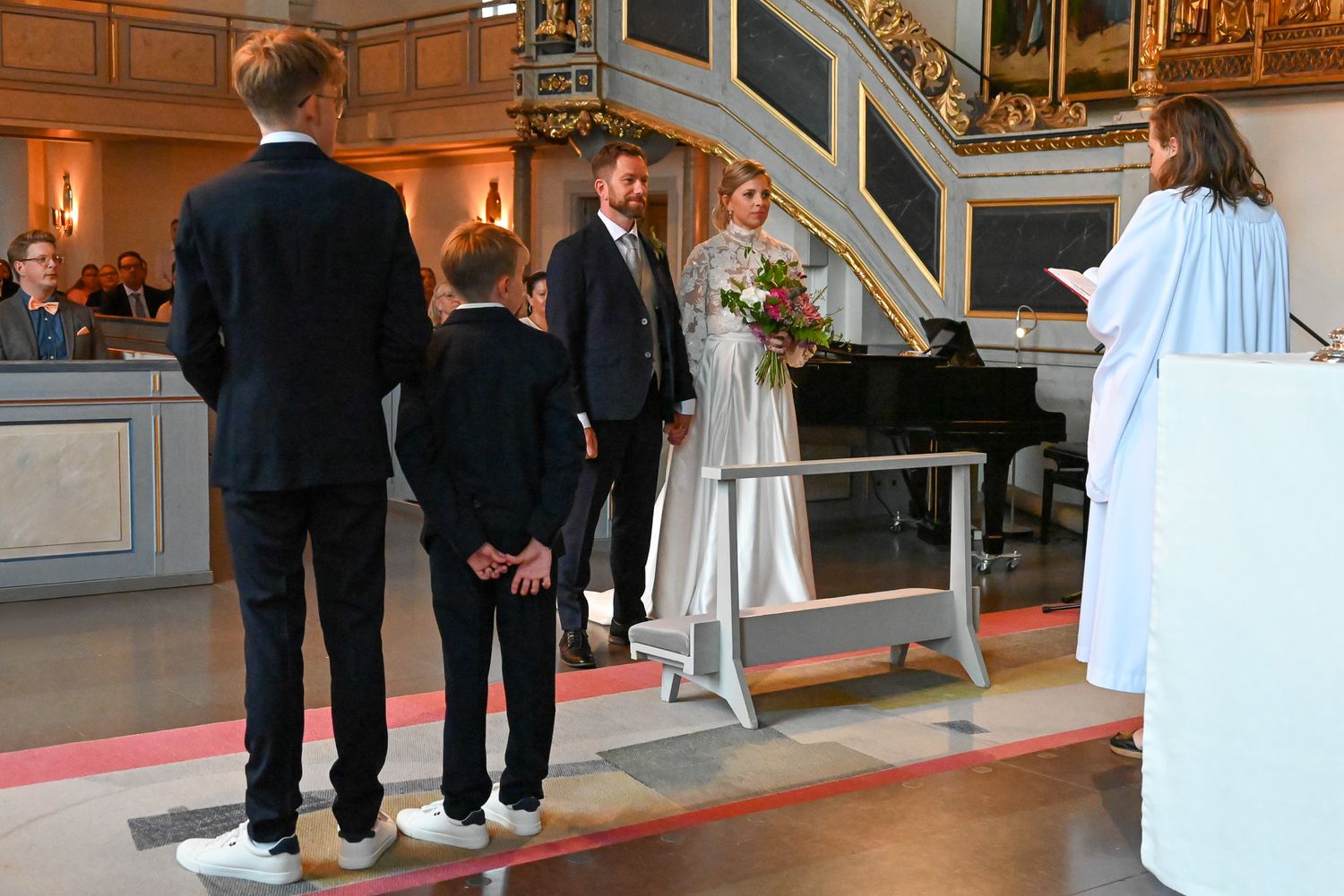 Foto inifrån vigselceremoni med brudparets barn och prästen framme vid altaret i detaljrik kyrka.