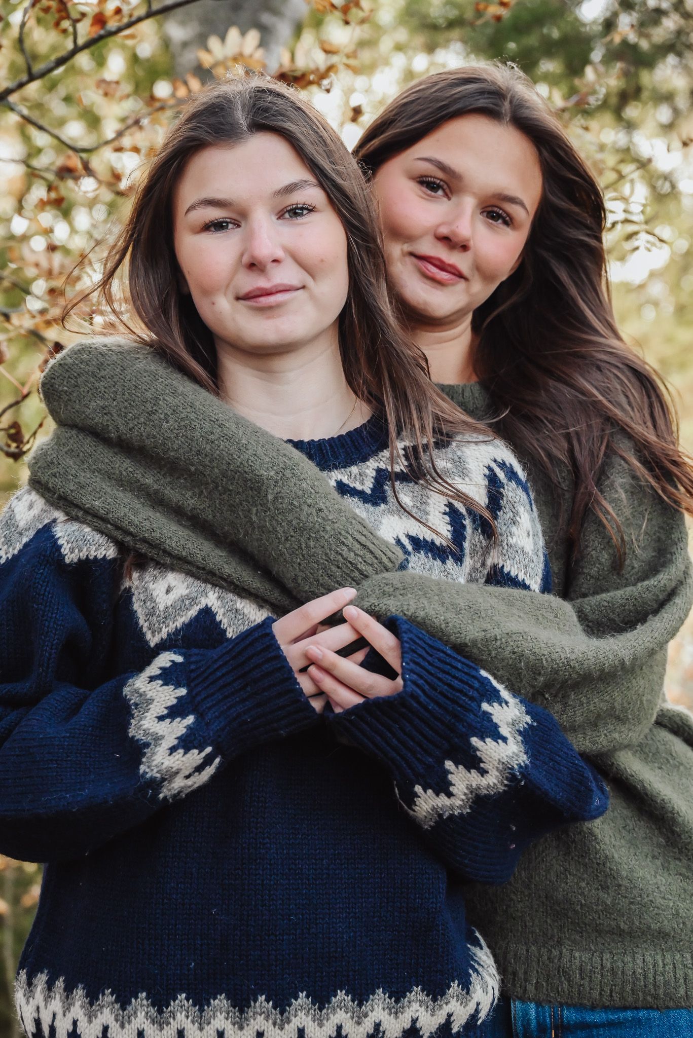 Två systrar håller om varandra i ett syskonfoto taget utomhus med höstfärger som bakgrund.