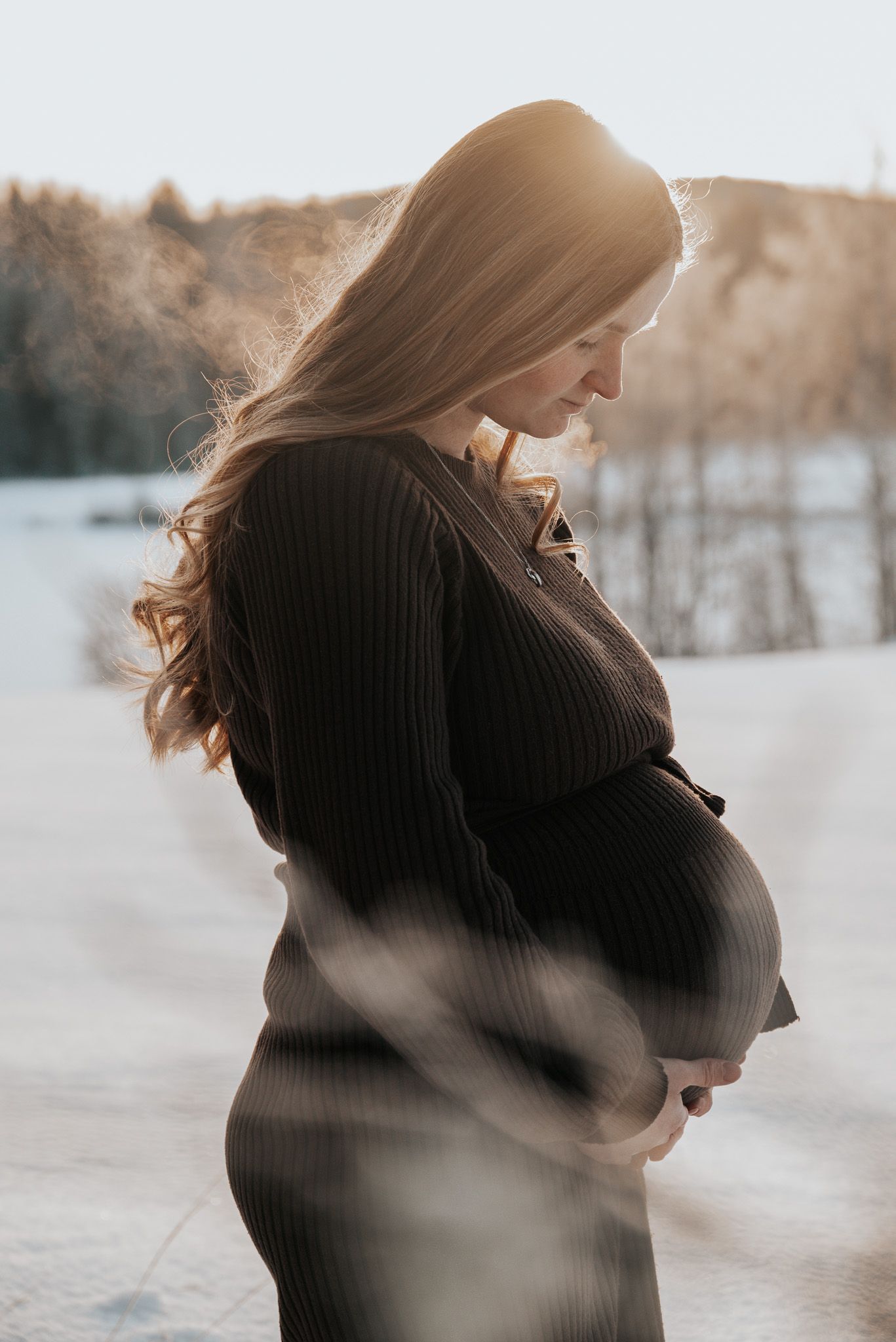 Gravid kvinna i mörk stickad klänning håller om magen kärleksfullt under golden hour i vinterlandskap.