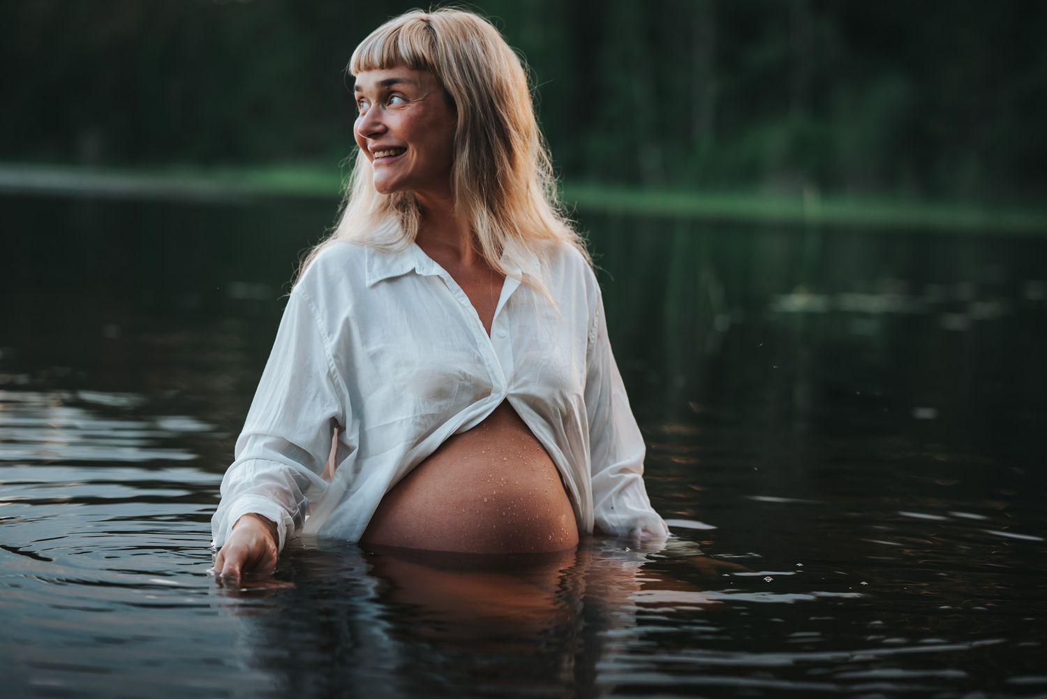 Leendes gravid kvinna i vit skjorta sitter i vattnet under gravidfotografering under golden hour med skog som bakgrund.