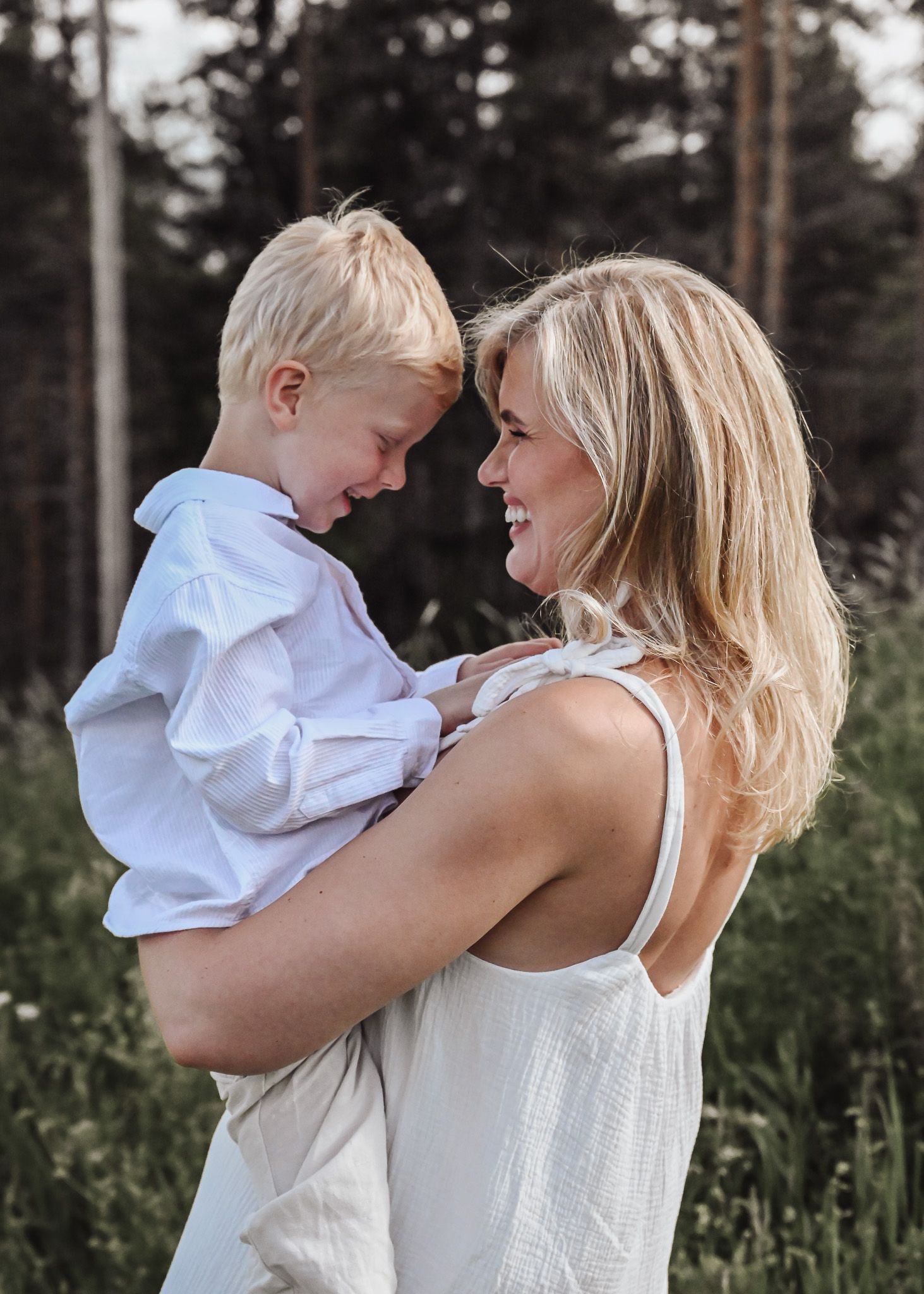 A mother and young child share tender moments outdoors in a forest setting wearing coordinating white outfits.
