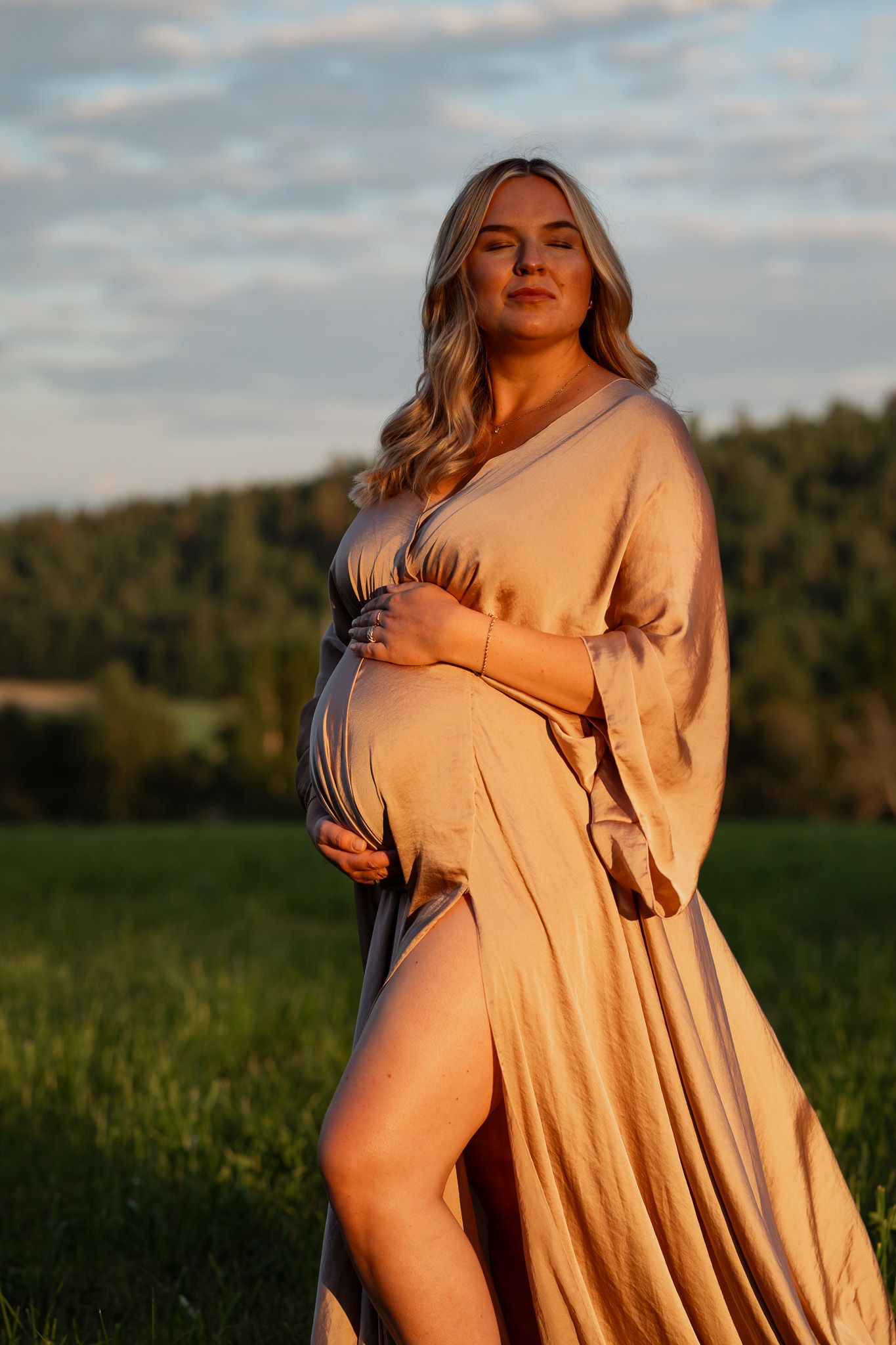 Gravid kvinna i beige gravidklänning som vinden tar tag i under golden hour med äng och skogslandskap i bakgrunden.