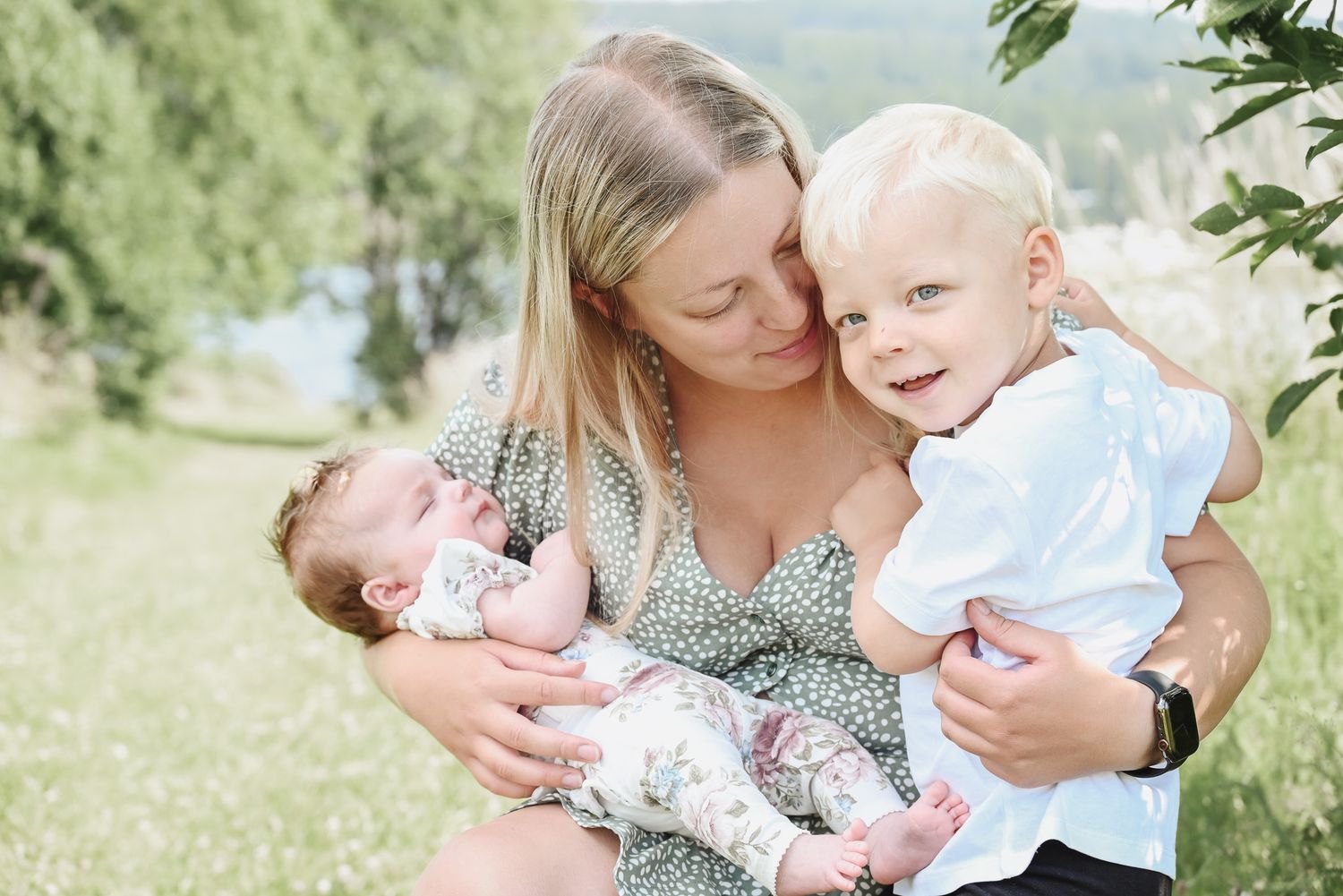 A loving family moment outdoors with a woman in a floral dress holding two young children on a sunny day.