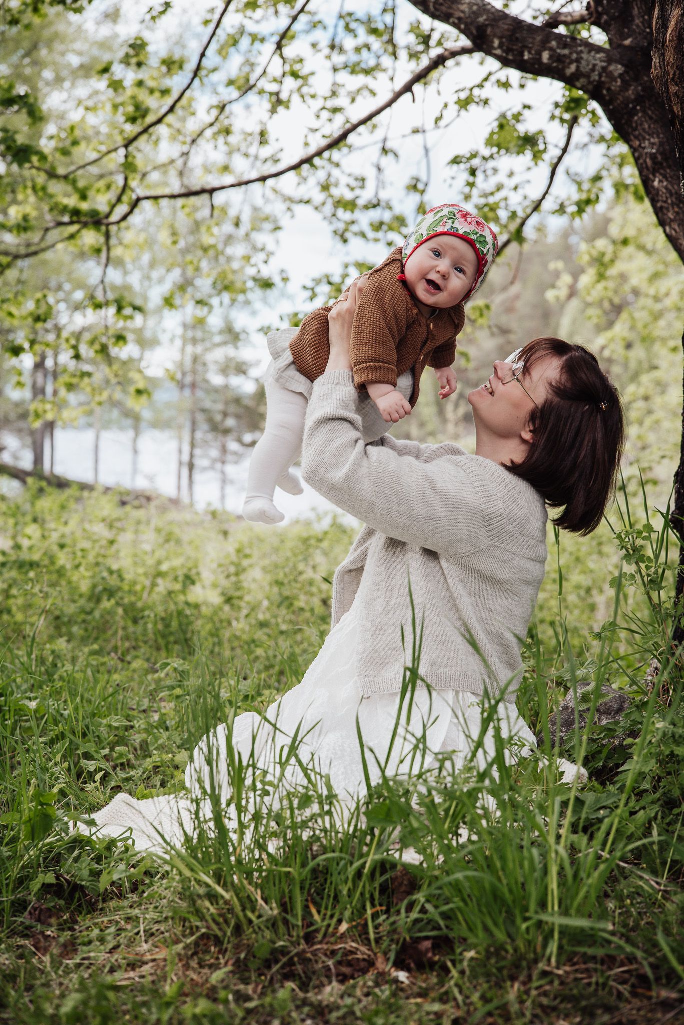 Mamma tillsammans med sin dotter sitter under stort träd i grönska och lyfter upp sitt skrattande barn.