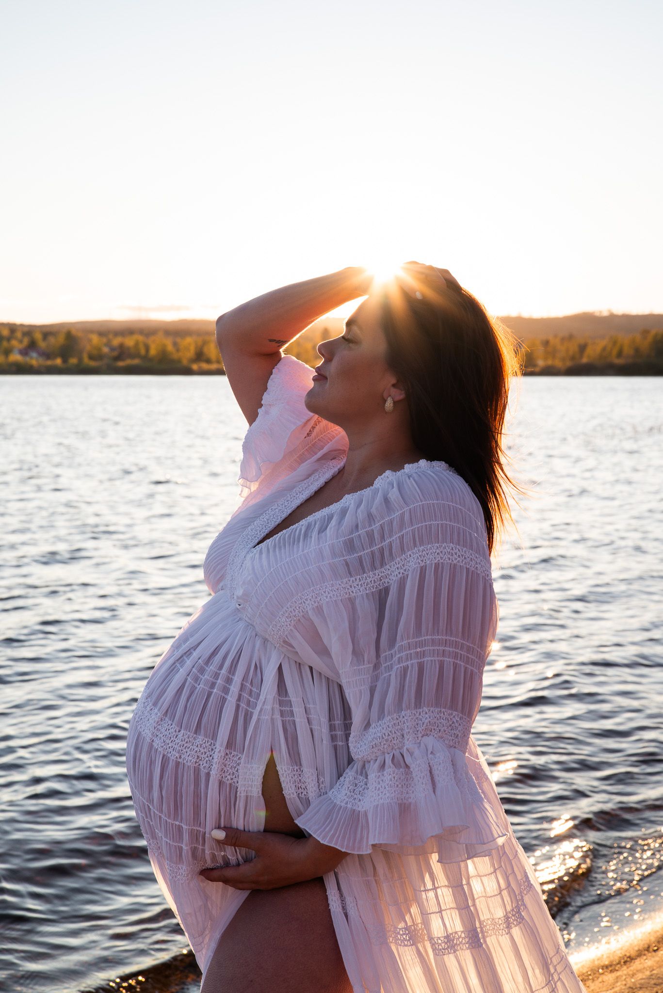 Gravid kvinna i spetsklänning står med handen i håret och håller om magen i solnedgången intill vattenbrynet.