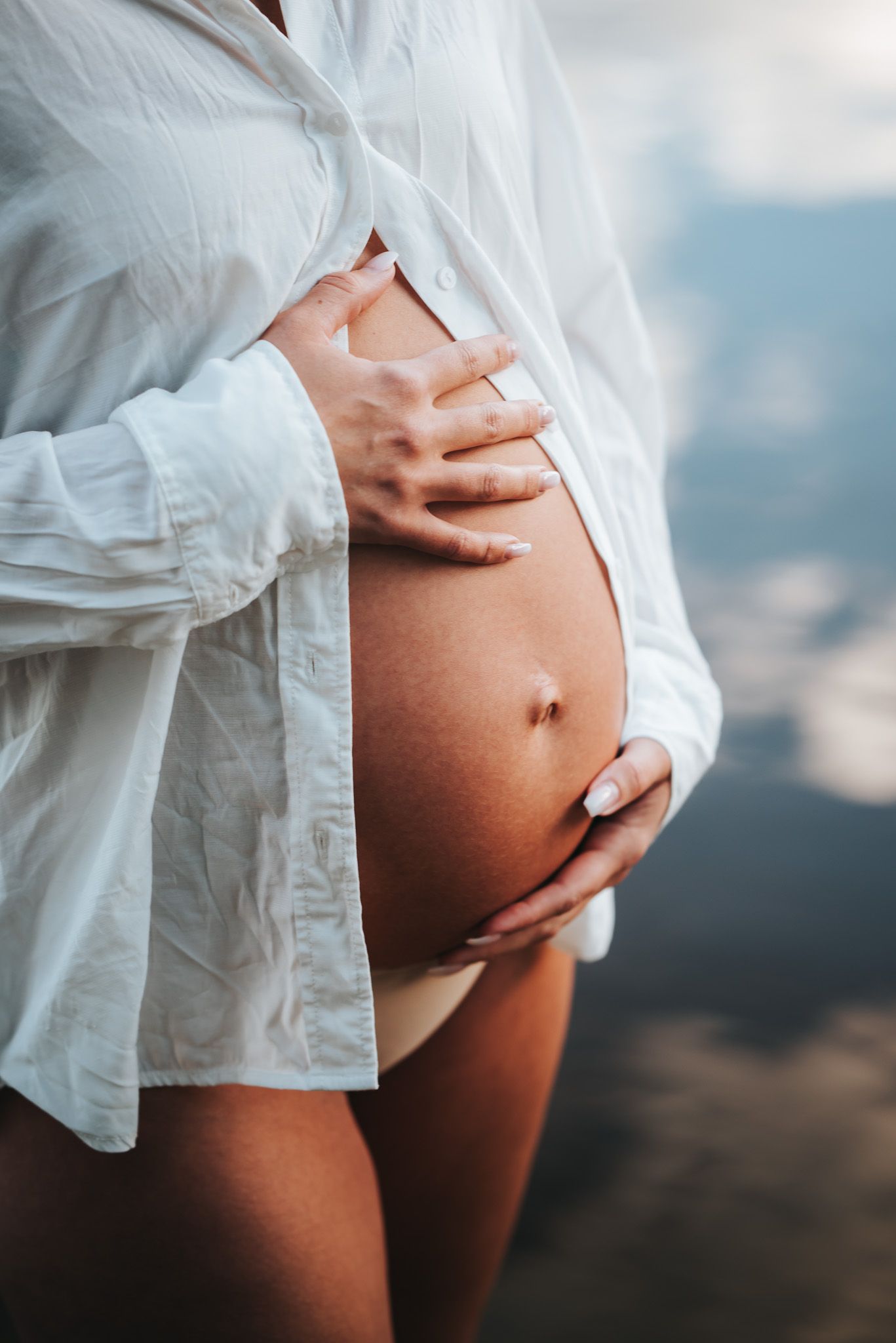 Gravid kvinna håller kärleksfullt om bebis i magen i vit skjorta i vattnet och med kvällssolen som lyser på magen och skapar mjukt ljus.