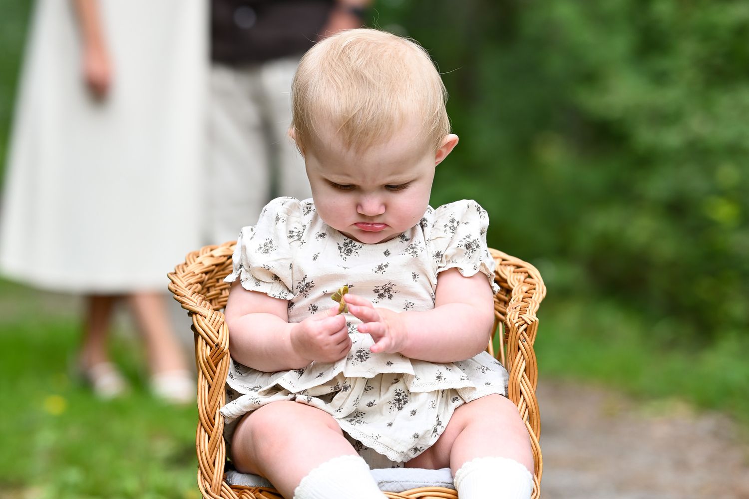 Bebis sitter i rottingstol och leker med ett löv medan föräldrar pussas i bakgrunden.