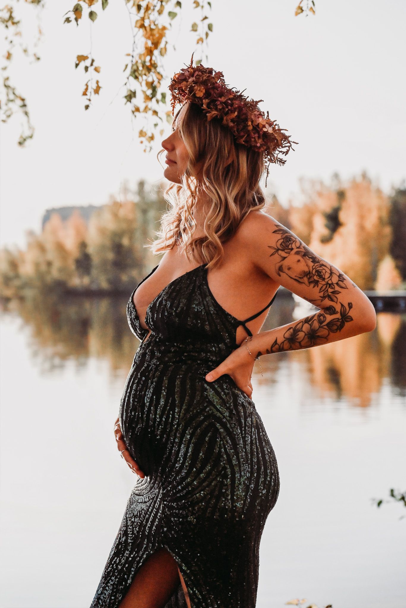 Gravid kvinna i grön tight klänning med glittrande paljetter och höstlöv i kransen står framför sjö med höstfärgade löv på träd.
