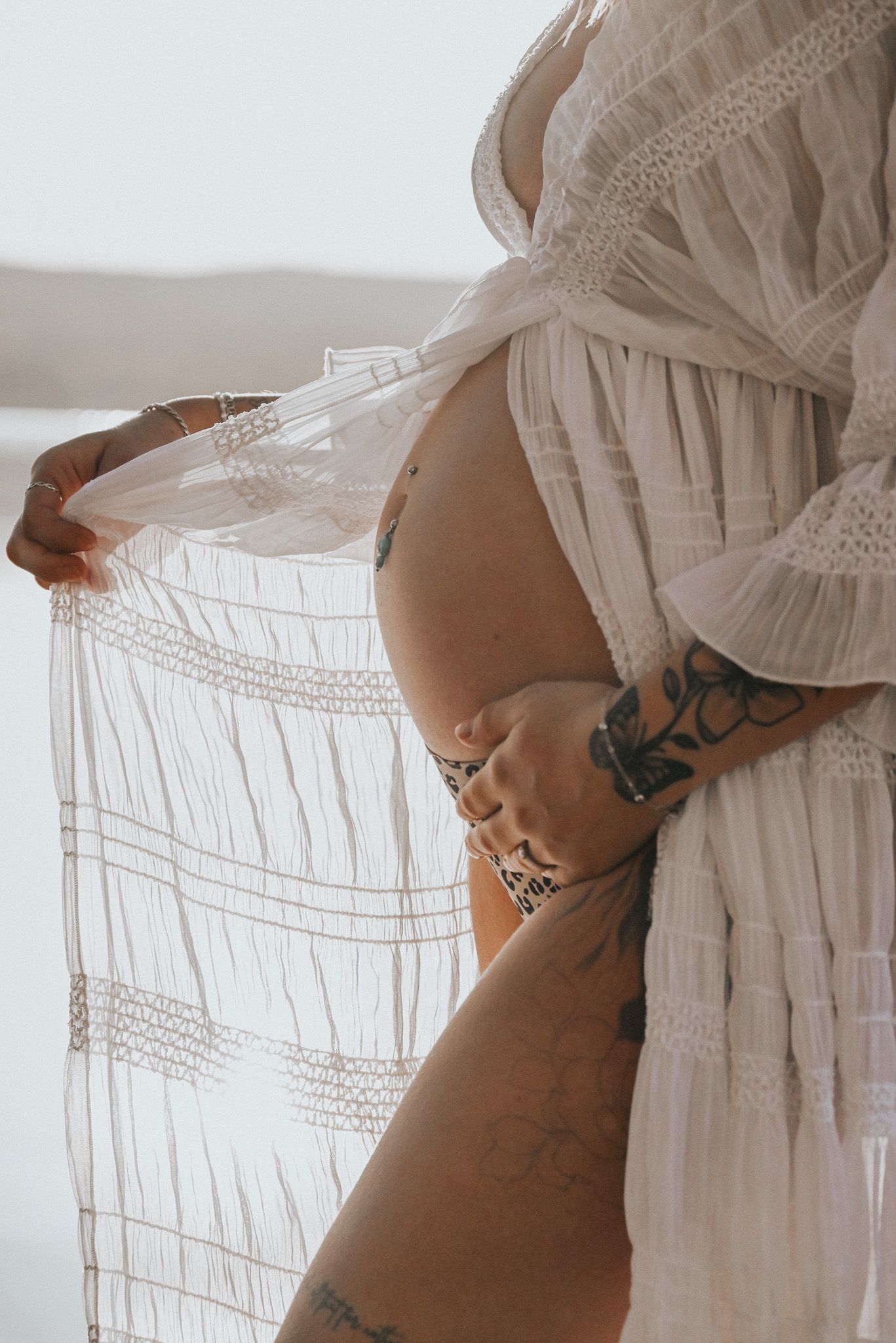 Smakfull gravidfotografering med detaljbild som visar gravidmage i profil mot vit spetsklänning och tatuerade armar som ger fint kontrast.