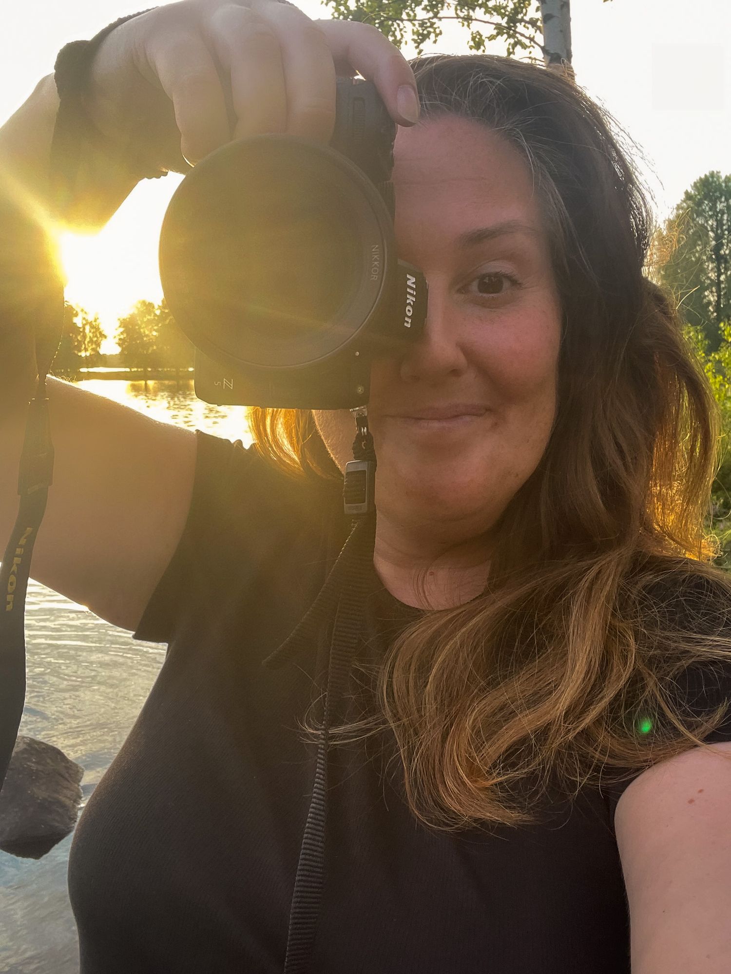 Selfie visar fotograf som ler i motljus under fotograferings session under golden hour intill sjö och skog.