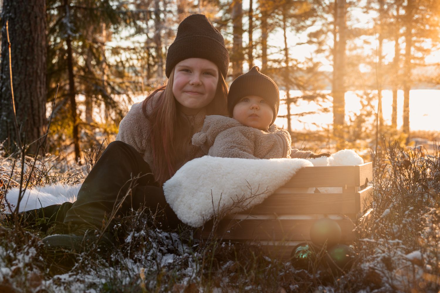 Lillasyster i låda med pälspläd blir kramad av storasyster under fotografering vintertid i motljus under golden hour.