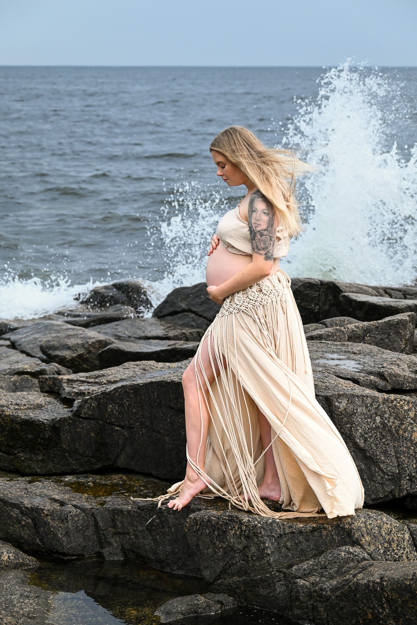 Gravid kvinna vid kusten med vågor som dramatiskt slår upp bakom henne under gravidfotografering i solnedgången.