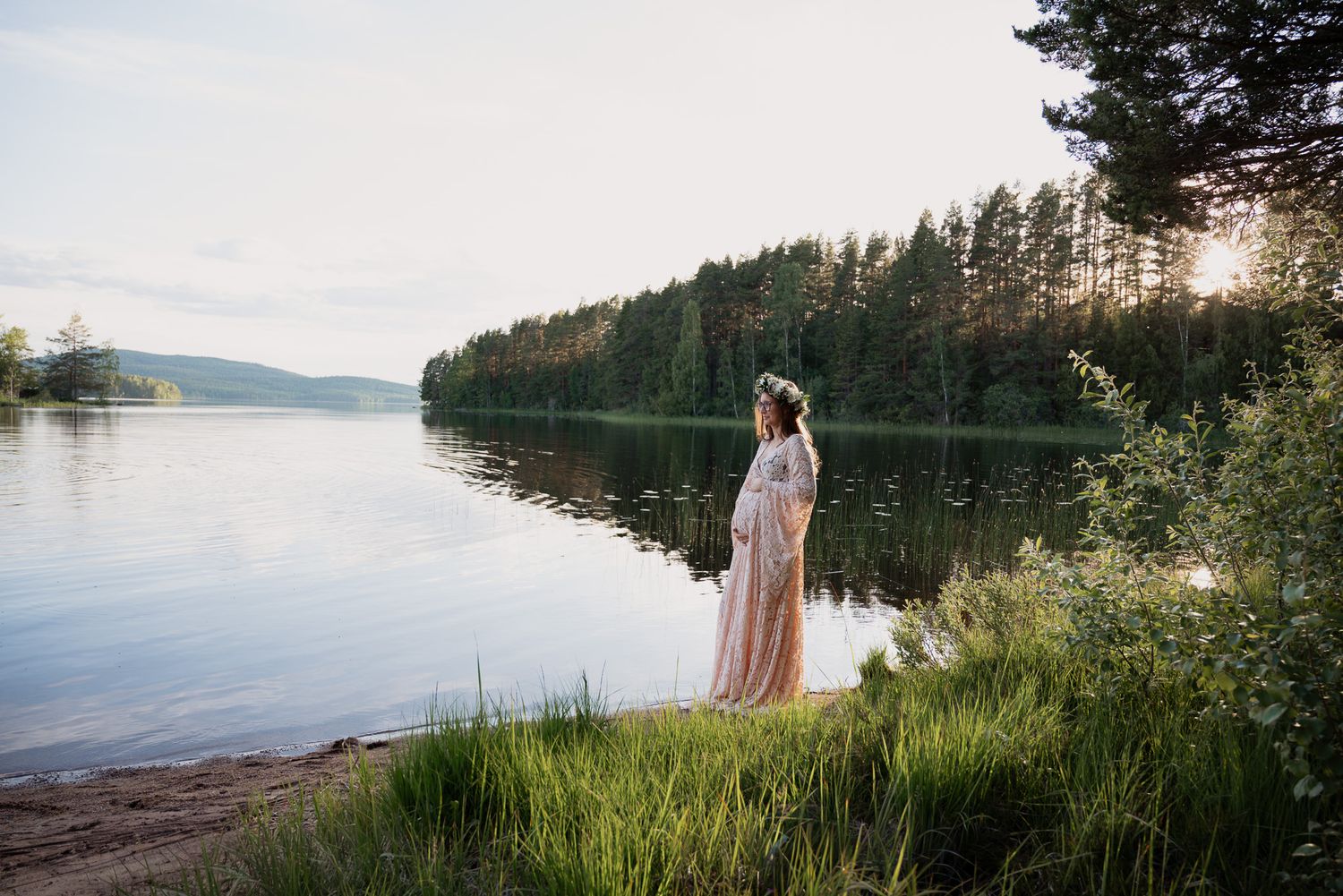 Gravid kvinna med somrig spetsklänning i vattenbrynet under golden hour.