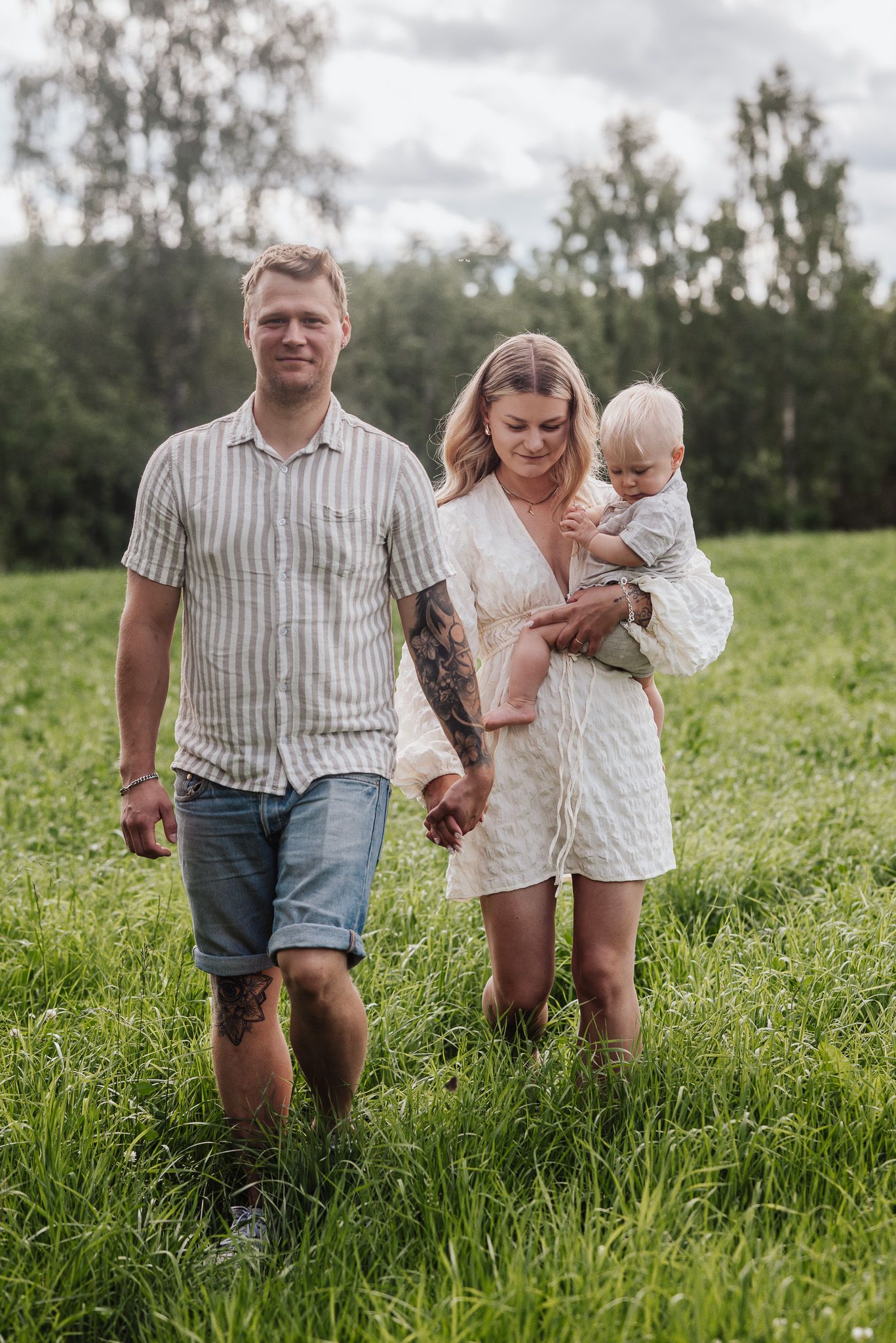 Familj går över äng tillsammans hållandes i hand och bärandes på sin son med träd och sommaräng i bakgrunden.