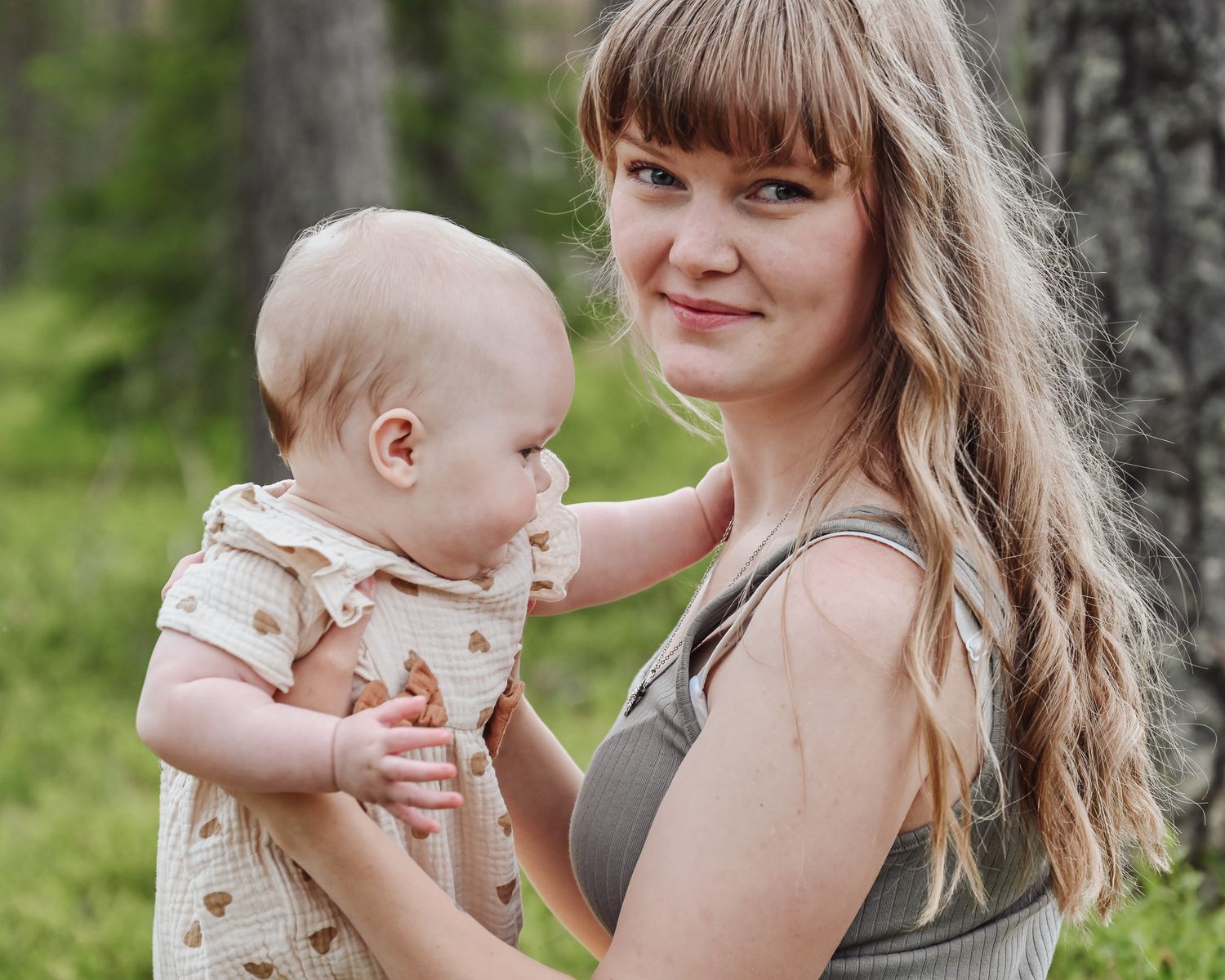 A parent holds a baby wearing a patterned onesie while standing outdoors with a green forest backdrop.