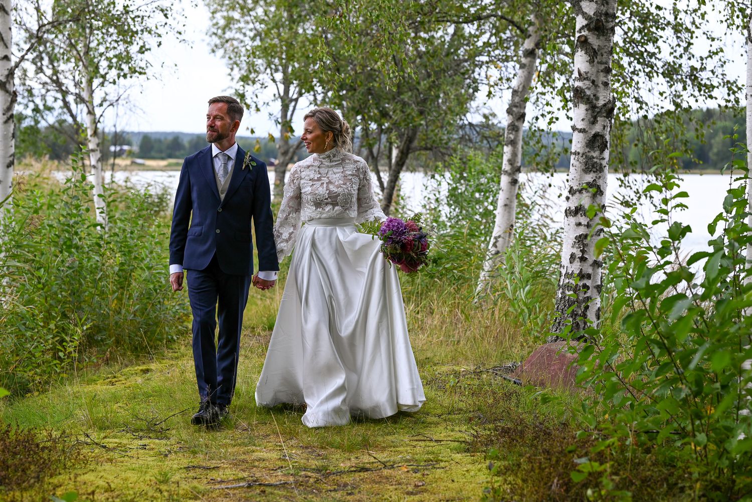 Brudpar strosar tillsammans hand i hand bredvid björkar intill sjö under bröllopsfotografering.