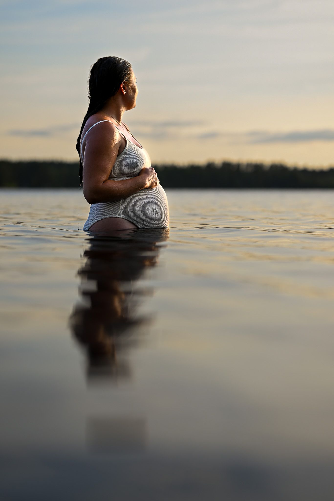 Gravid kvinna står i vatten under golden hour med händerna på gravidmagen och reflektionen från henne som syns på vattenytan.